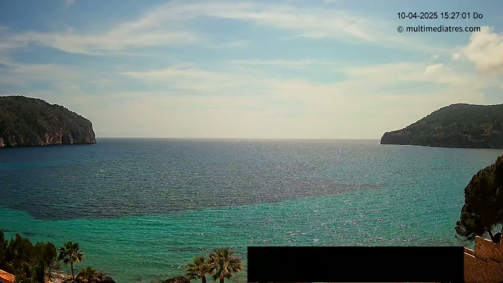 Under a partly cloudy sky, a tranquil bay with teal water is nestled between two dark, rocky hills.