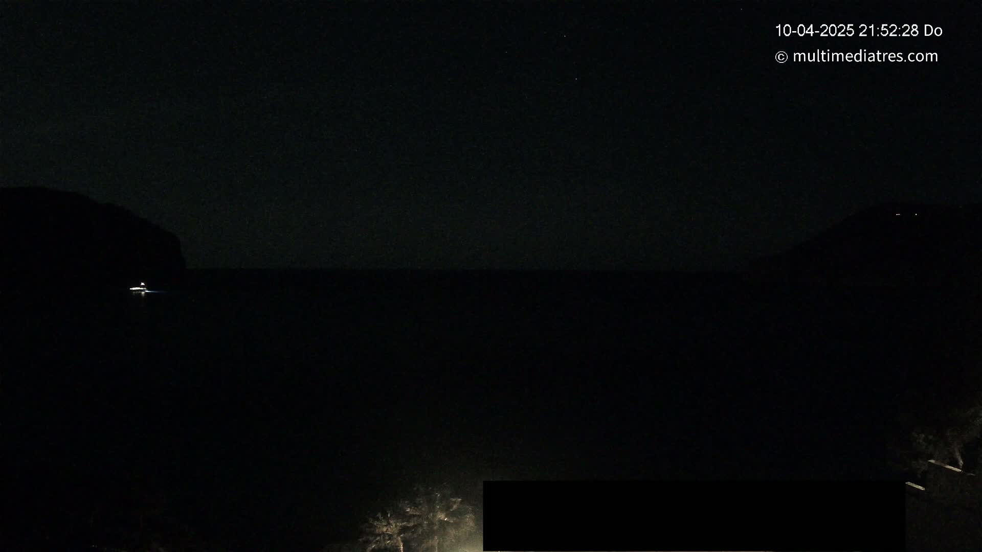 A dark nighttime scene shows a body of water with a small light in the distance, silhouetted landmasses on either side, and dark vegetation in the foreground under a clear, starlit sky.