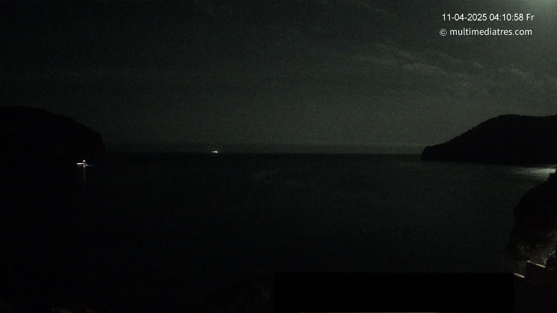A nighttime view of a dark body of water between two silhouetted hills under a mostly cloudy sky.
