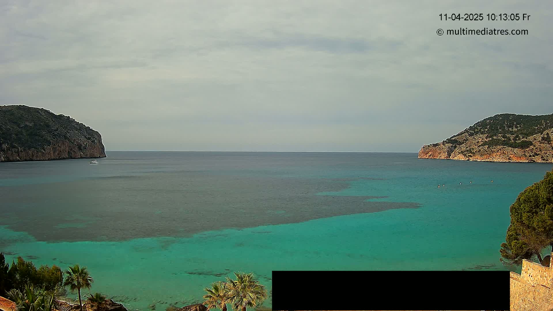 A calm, overcast day reveals a picturesque bay with teal water, flanked by rocky hills, and a few small boats visible in the distance.