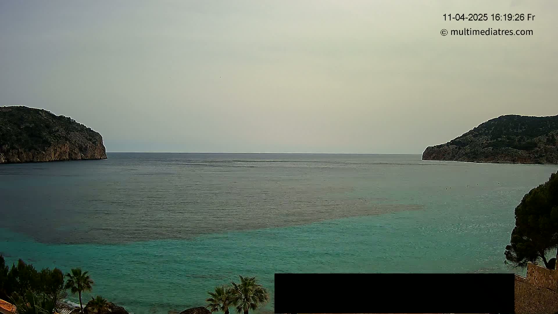 A calm, overcast day shows a bay of teal water between two dark, rocky hillsides, with vegetation along the shore in the foreground.