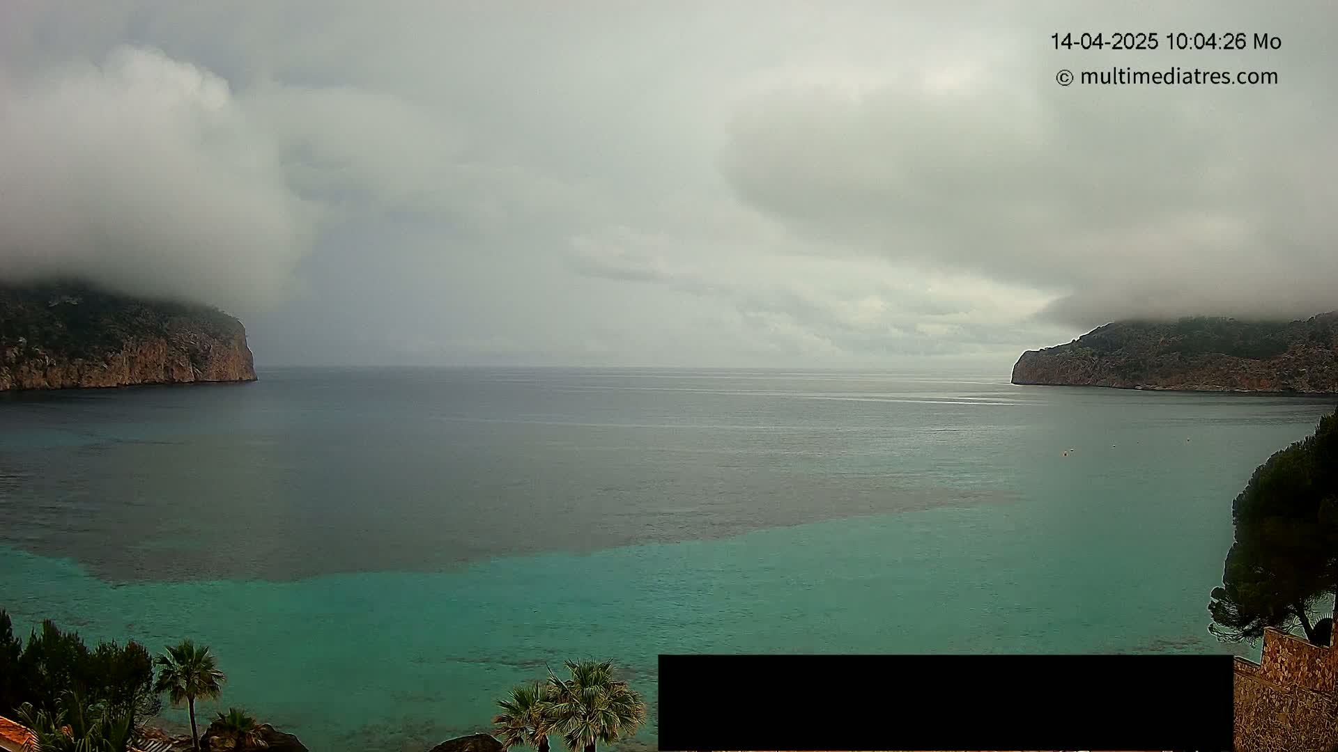 A partly cloudy, overcast day reveals a tranquil bay with teal water between two rocky headlands, one of which is shrouded in fog.
