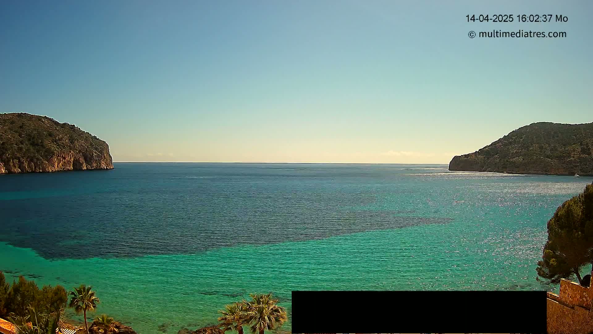A clear, sunny day reveals a calm, turquoise bay nestled between two rocky headlands.