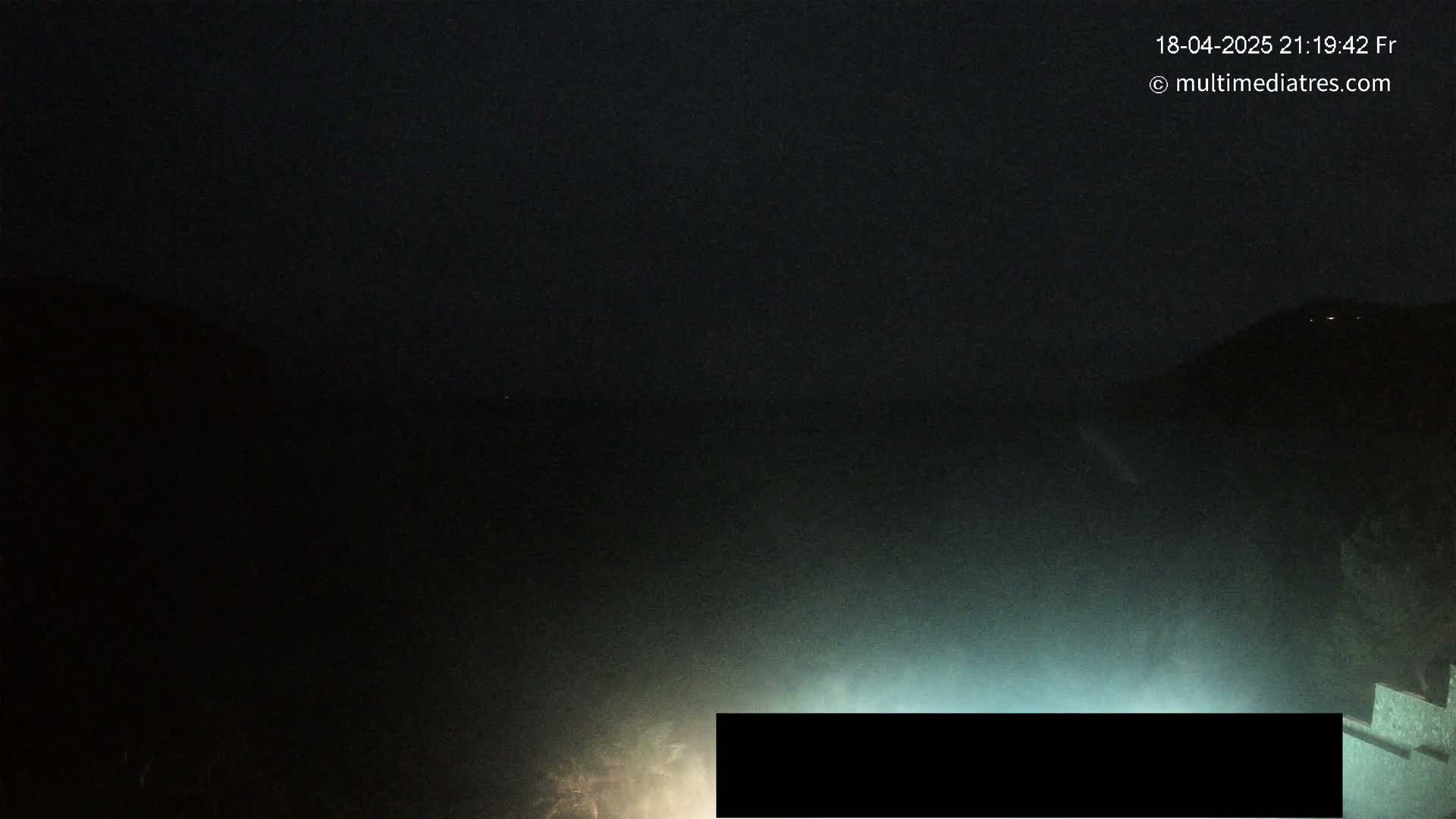 A nighttime, hazy, outdoor scene shows dark hills and a low light source near the bottom of the frame.