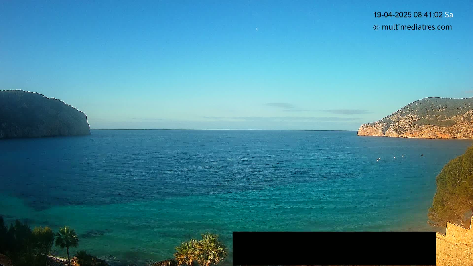 A calm, clear blue ocean is nestled between two rocky hills under a bright, sunny sky.