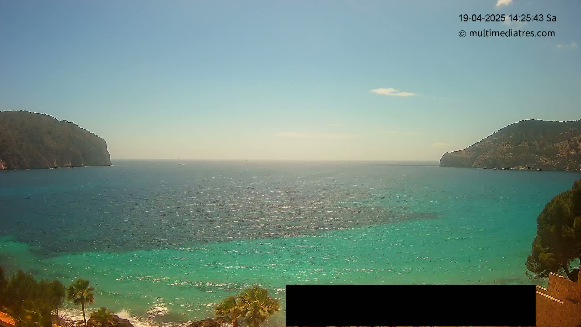 A clear, sunny day reveals a tranquil bay with turquoise water, bordered by dark, rocky hillsides and dotted with palm trees near the shore.