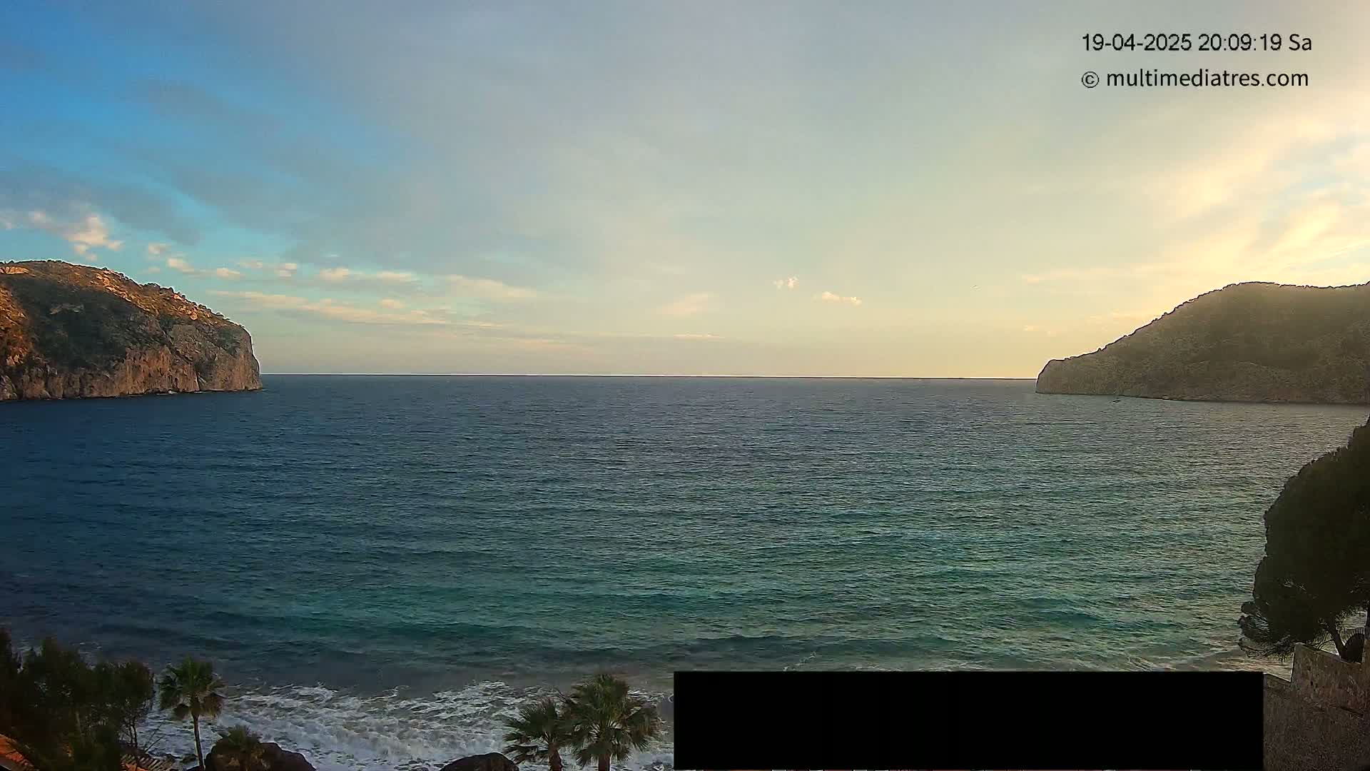 A calm sea with small waves laps against a shore lined with palm trees and vegetation under a partly cloudy, twilight sky, with two rocky cliffs visible in the distance.
