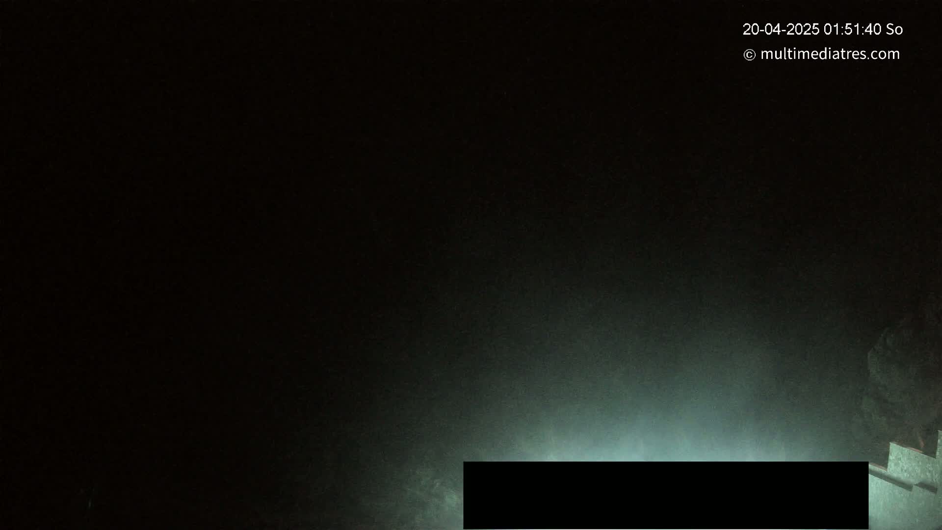 A low-light, nighttime image shows a dark, possibly foggy or misty scene with a faintly illuminated, rectangular object at the bottom.