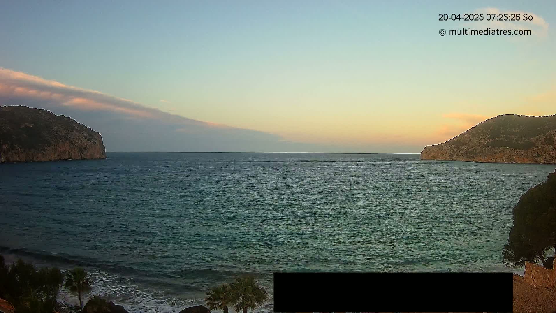 A calm sea at sunset is nestled between two rocky hills, under a mostly clear sky with a thin layer of clouds.