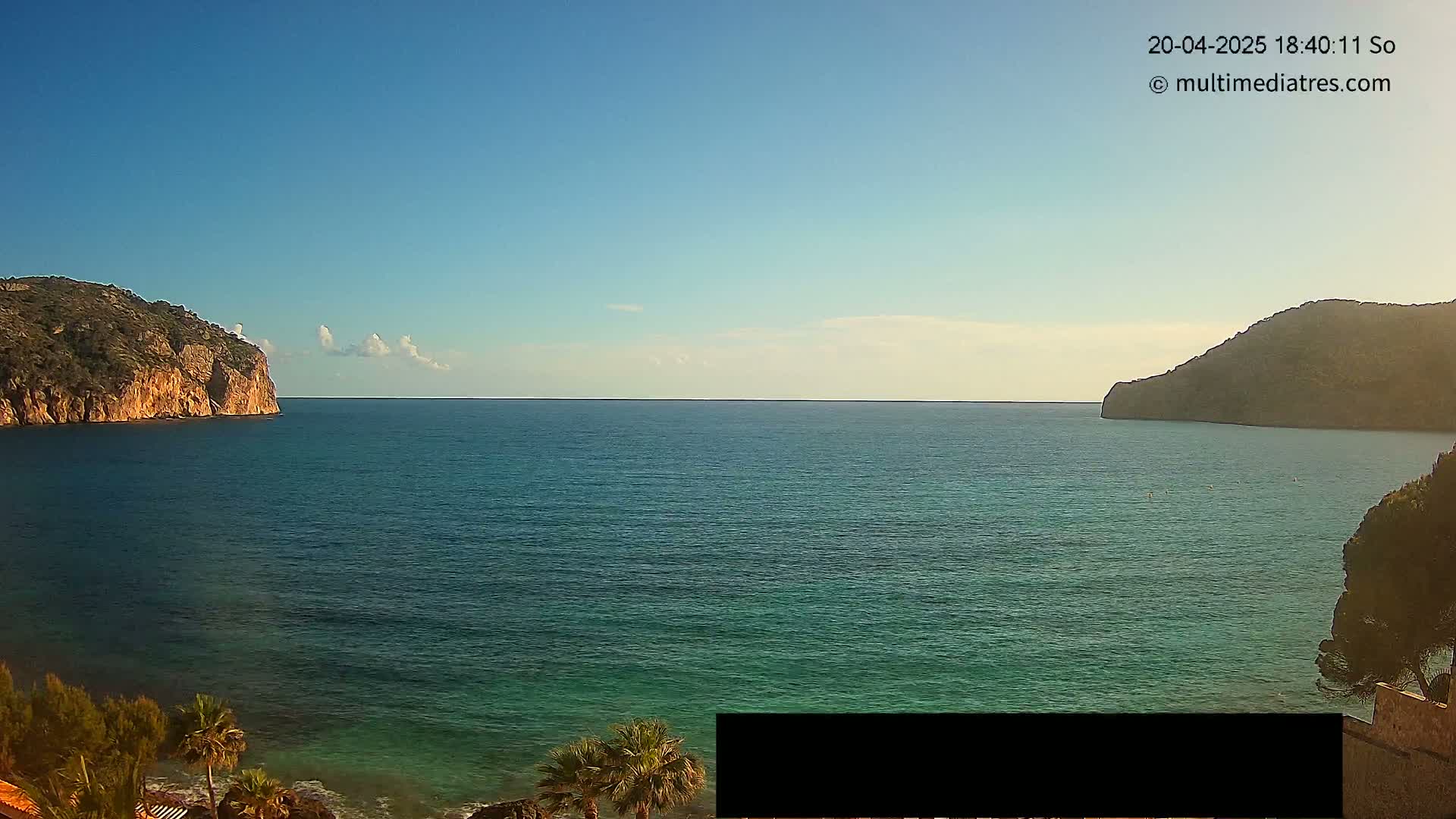 A calm, blue sea separates two rocky hills under a clear, sunny sky.