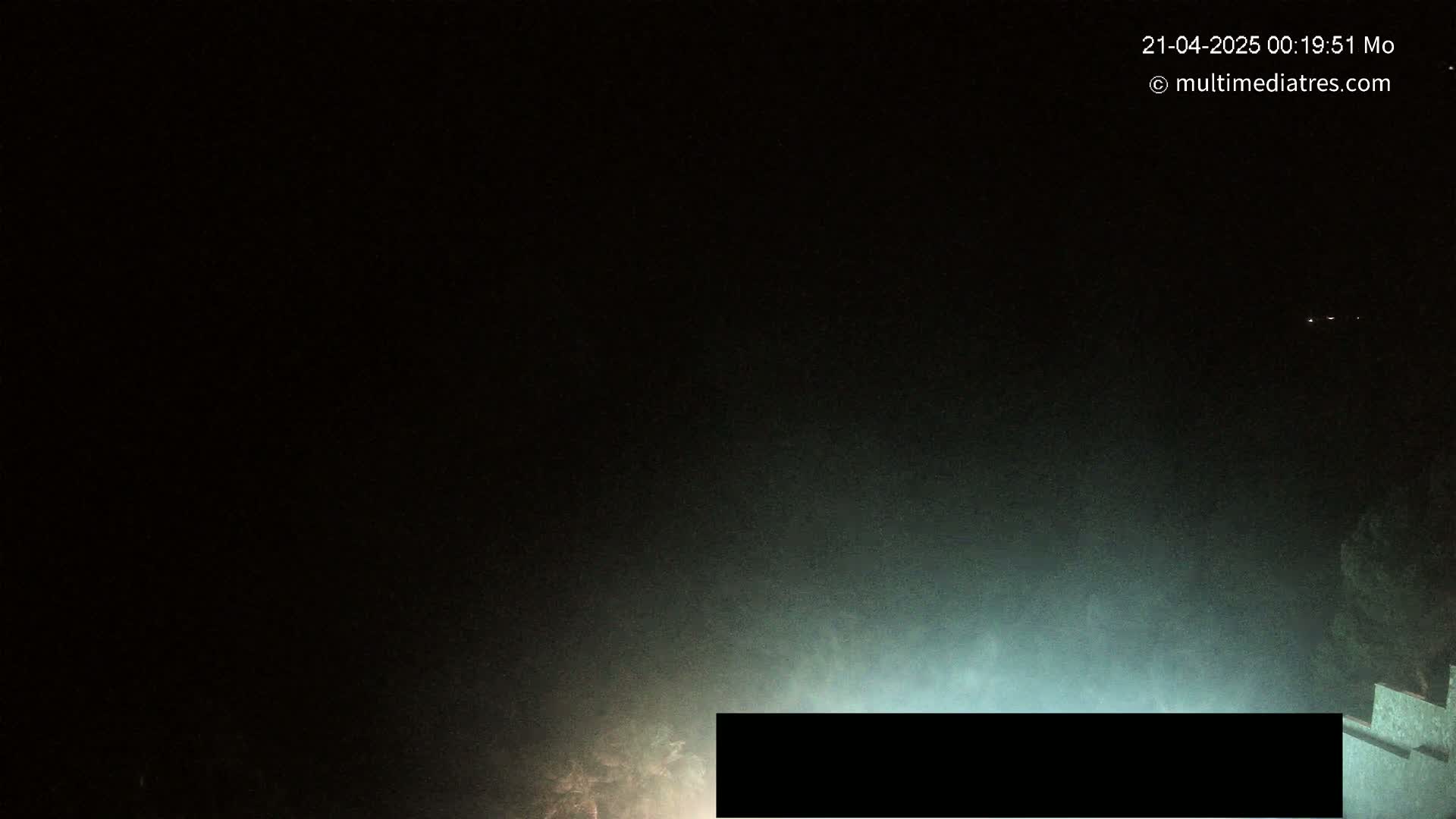 A nighttime, hazy outdoor scene shows a dimly lit structure partially obscured by fog or mist.