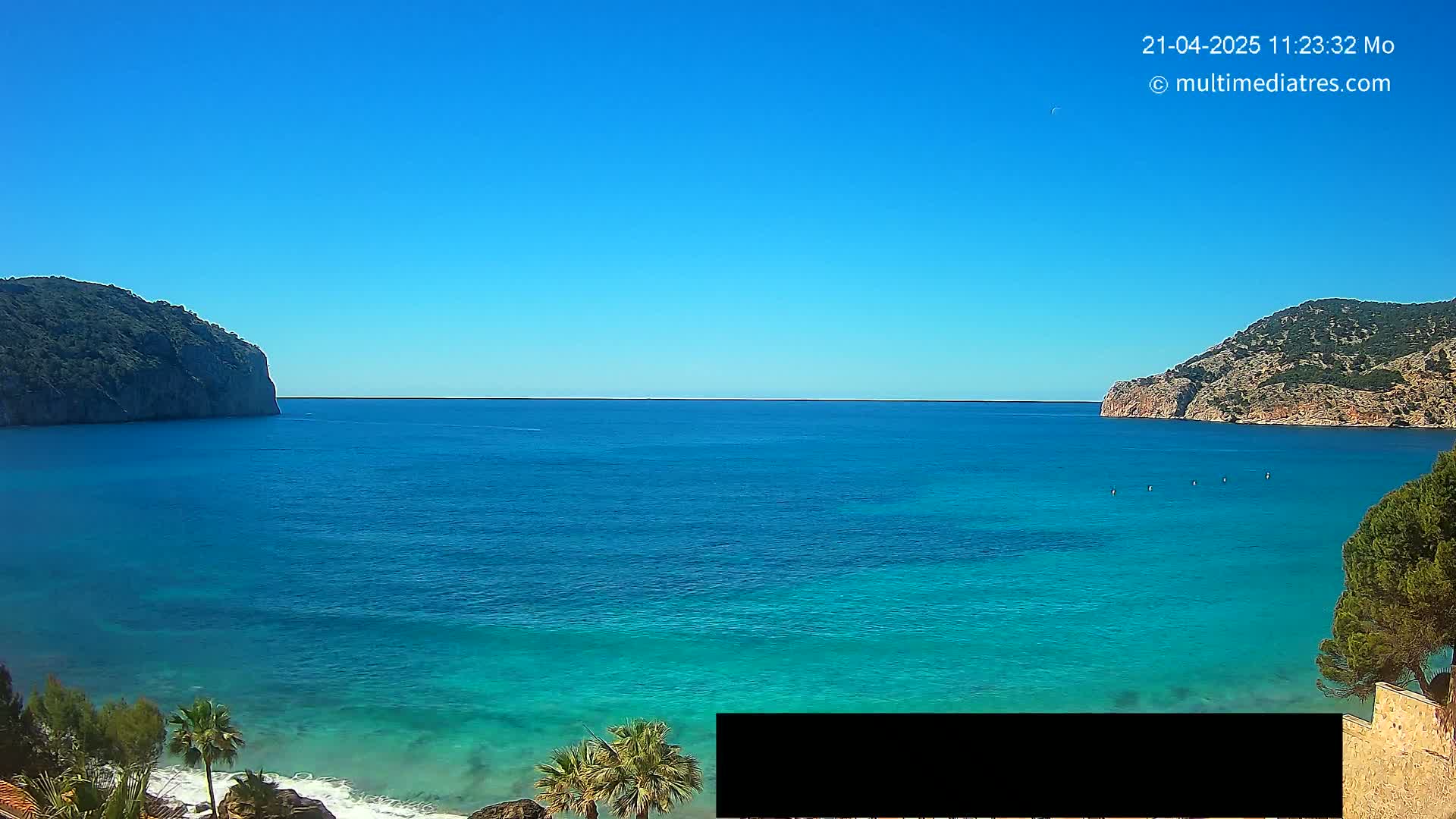 A clear blue sky overlooks a calm, turquoise ocean bay nestled between two rocky hillsides, with a few palm trees and some vegetation visible in the foreground.