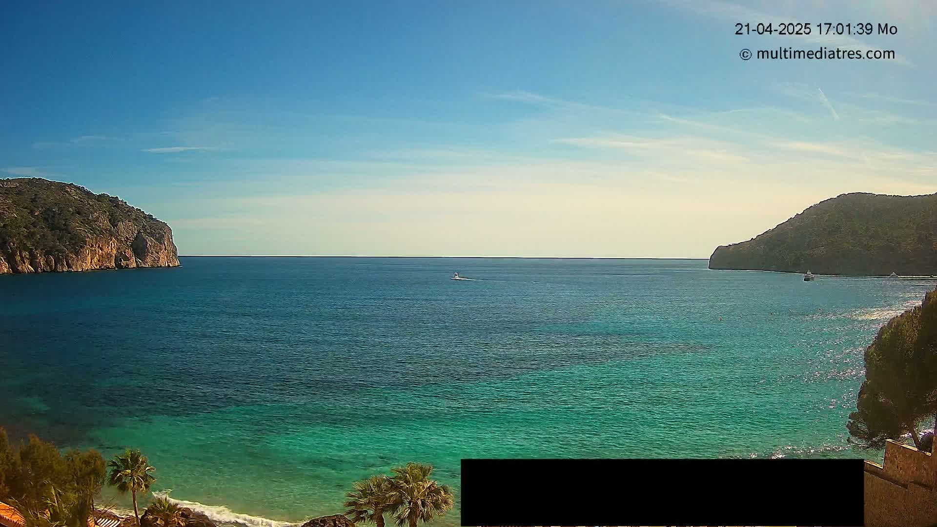 A calm, clear day reveals a picturesque coastal scene with teal-colored water nestled between two rocky hills, a small boat visible in the distance.