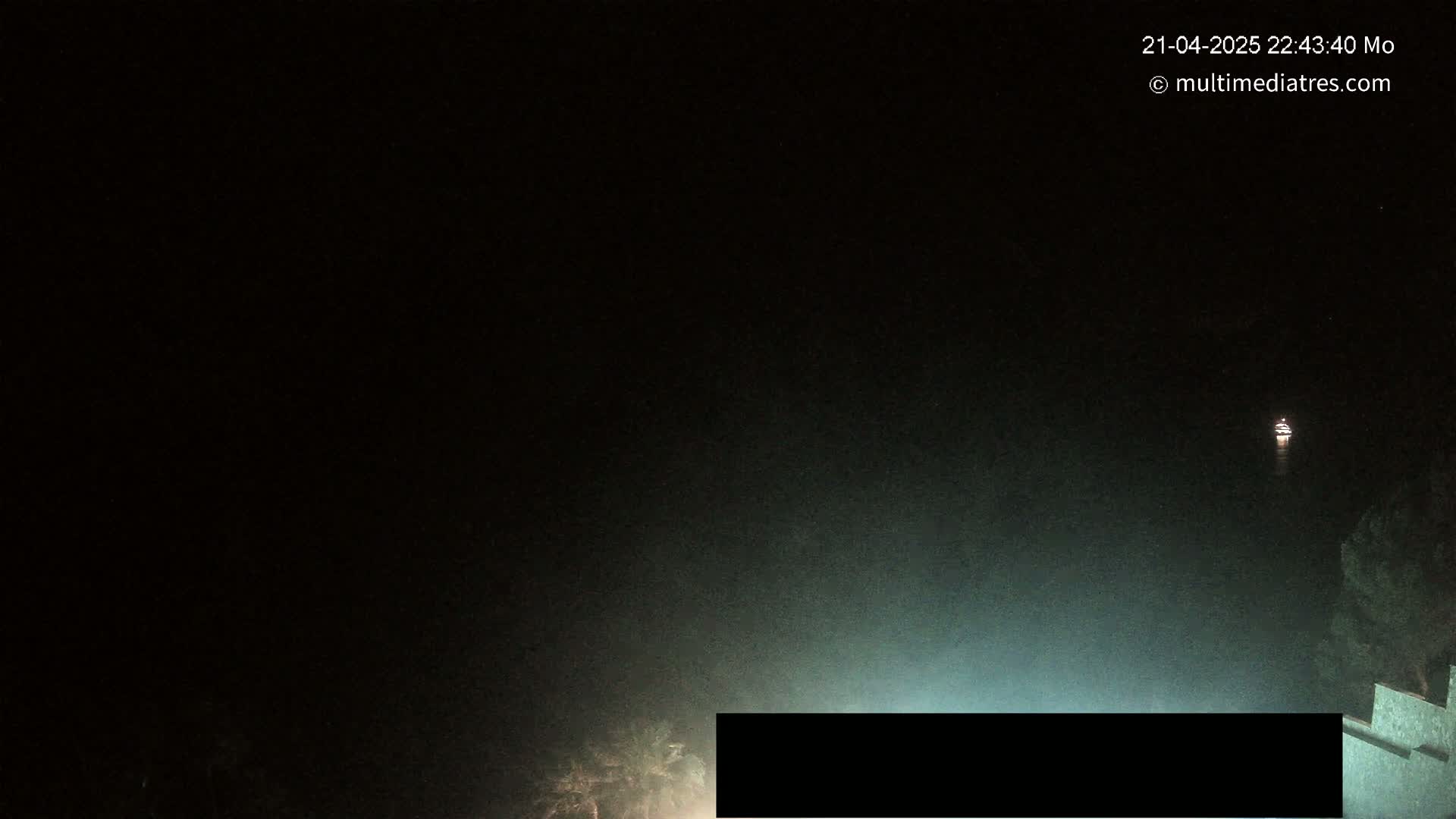 A dark, foggy nighttime scene shows a distant light on the water and a portion of a structure in the bottom right corner.