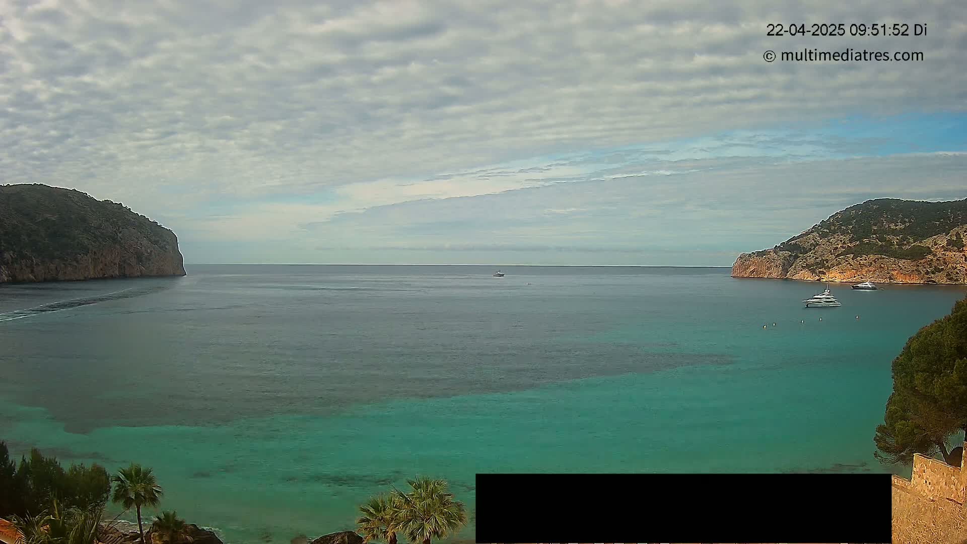 A tranquil bay with clear, teal water is nestled between two rocky cliffs under a partly cloudy sky.