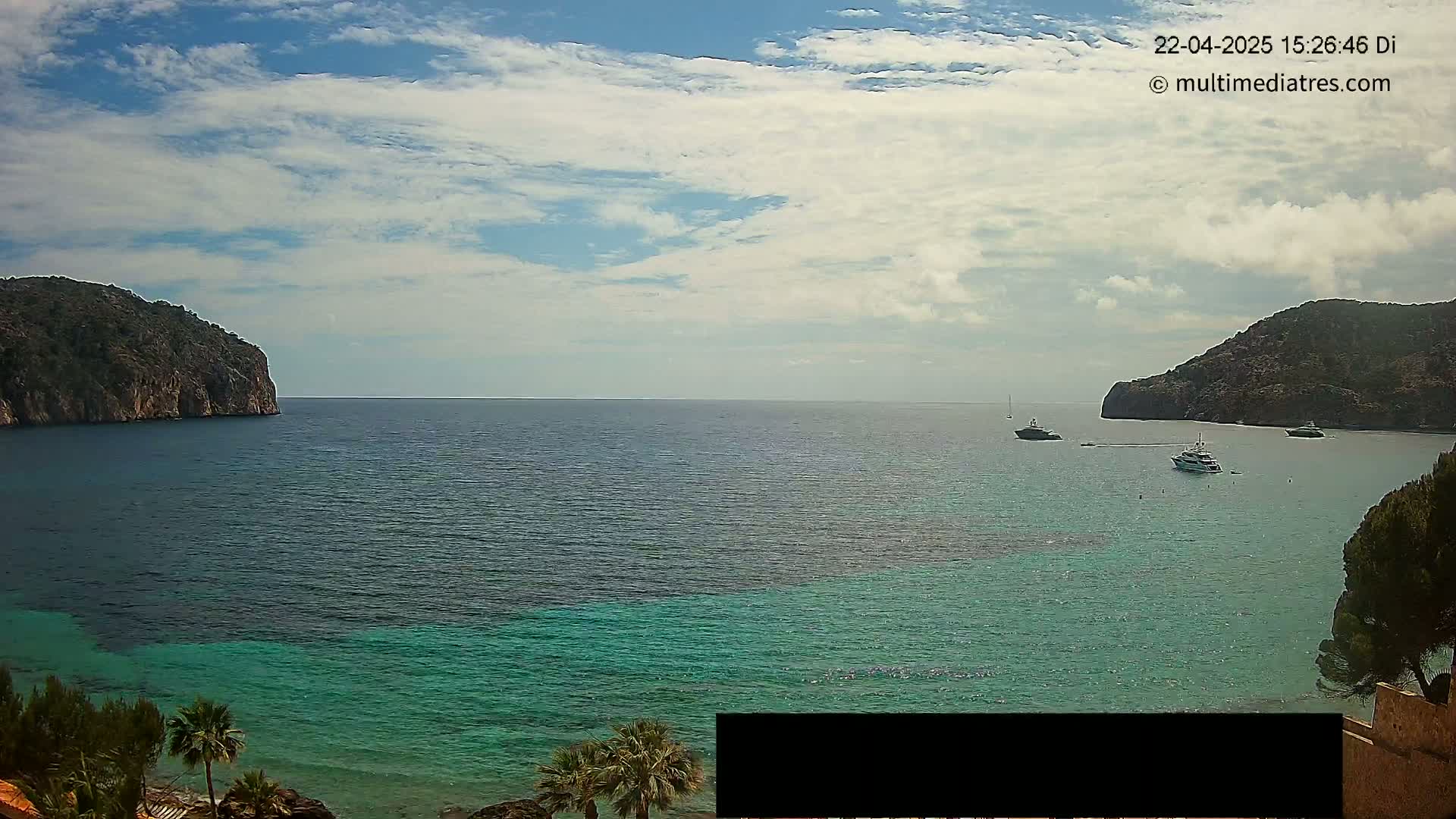 A partly cloudy day shows a calm bay of teal and deep blue water, nestled between two rocky hills, with several boats at anchor.
