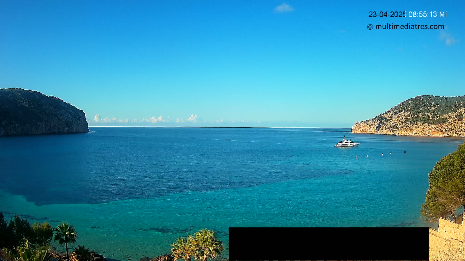 A yacht is visible on a calm, clear blue sea, enclosed by two rocky hills under a mostly clear sky with a thin line of clouds on the horizon.