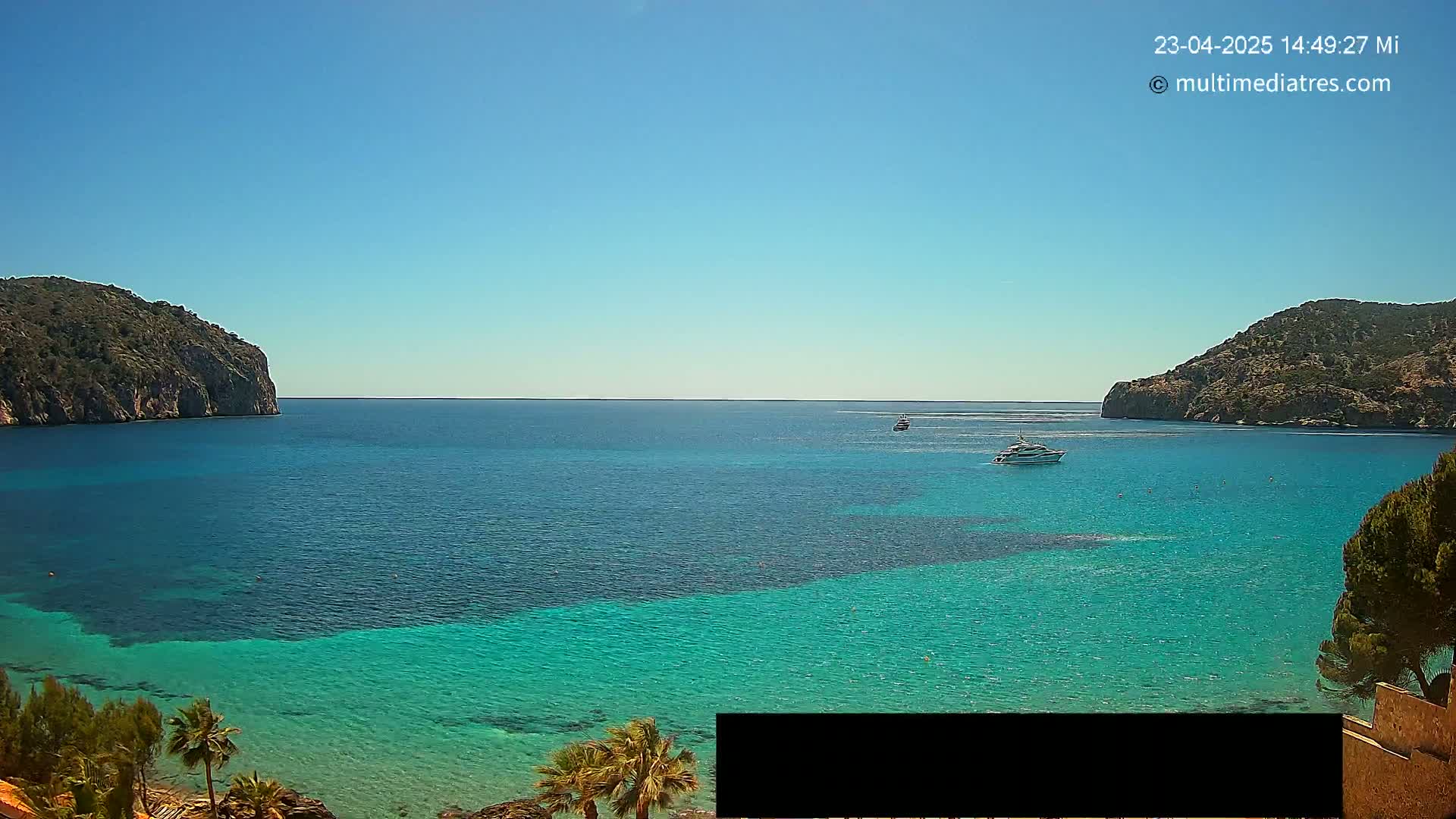 A clear, sunny day reveals a picturesque bay with turquoise water, two boats, and rocky hillsides on either side.