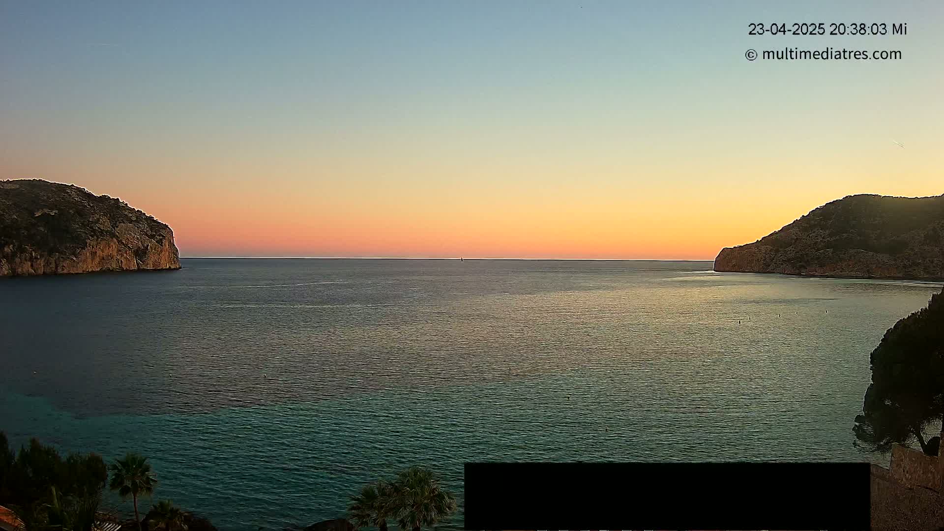 A calm sea at sunset is nestled between two rocky hills, with palm trees visible in the foreground.