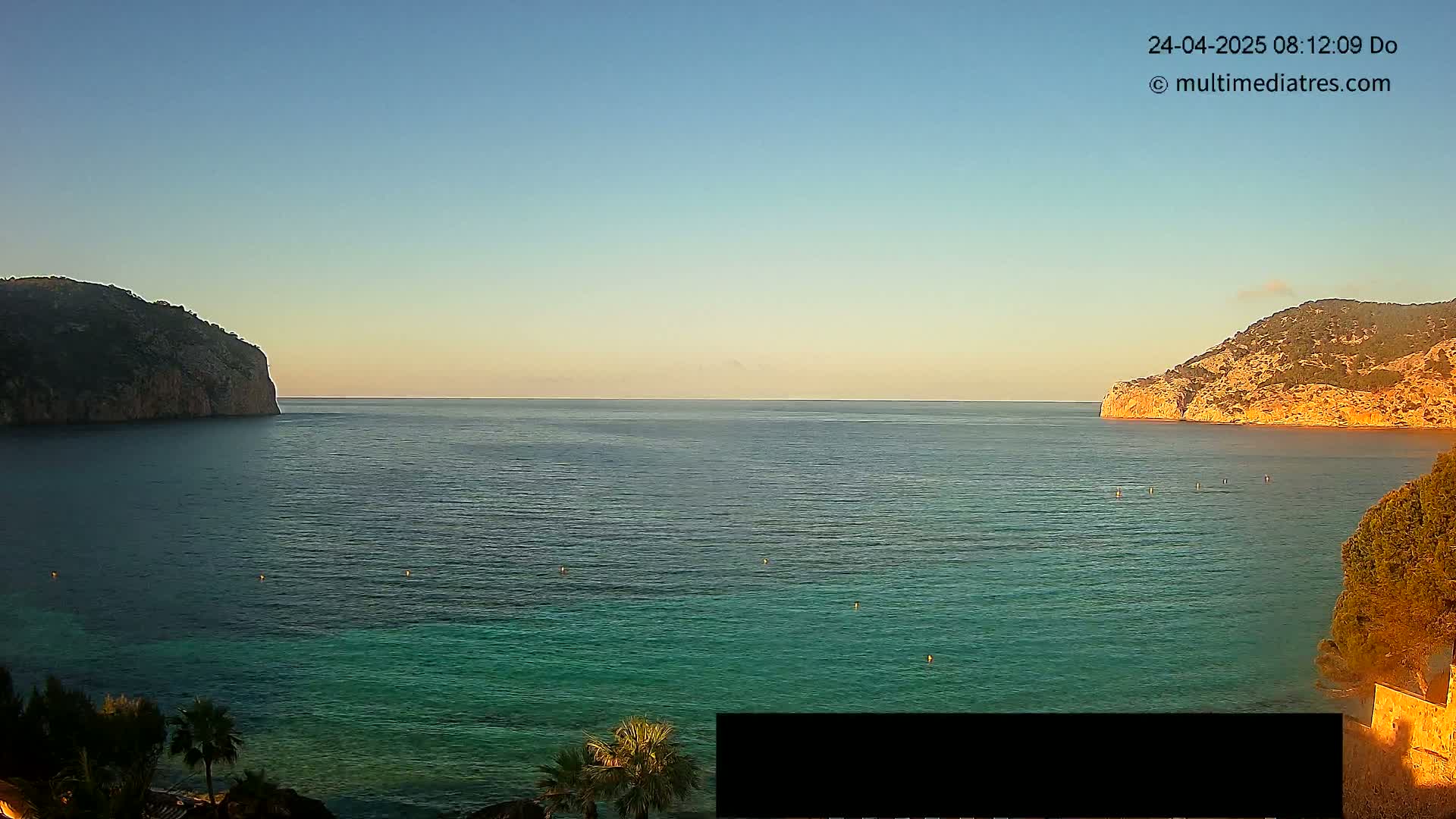 Under a clear, pale blue sky, a calm, teal sea stretches between two rocky, sparsely vegetated headlands.