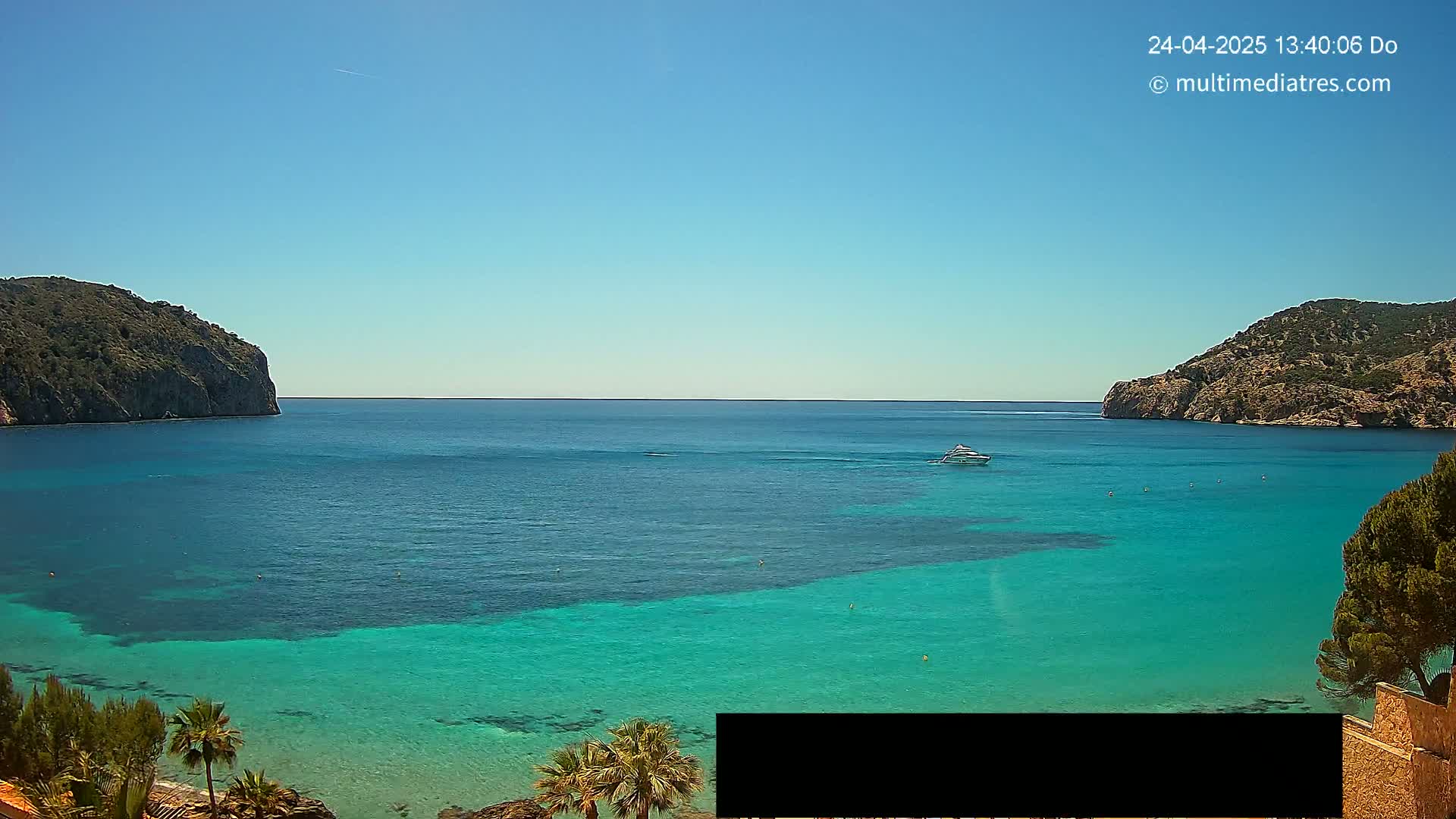 A clear, sunny day reveals a tranquil bay with vibrant turquoise water, flanked by two rocky hillsides and a lone boat on the calm sea.
