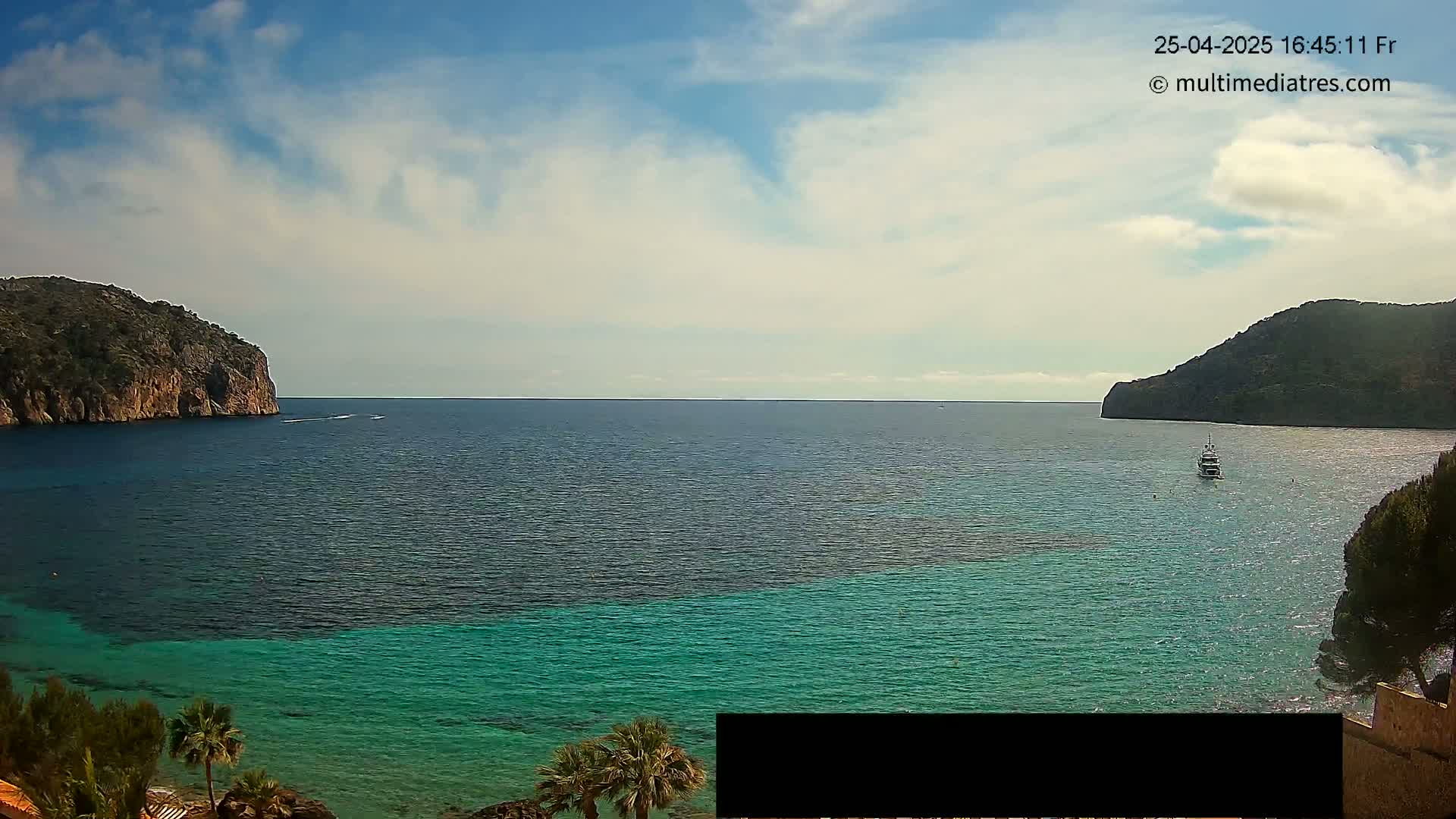A partly cloudy sky overlooks a calm bay of teal and dark blue water, with two rocky hillsides on either side and a boat in the distance.