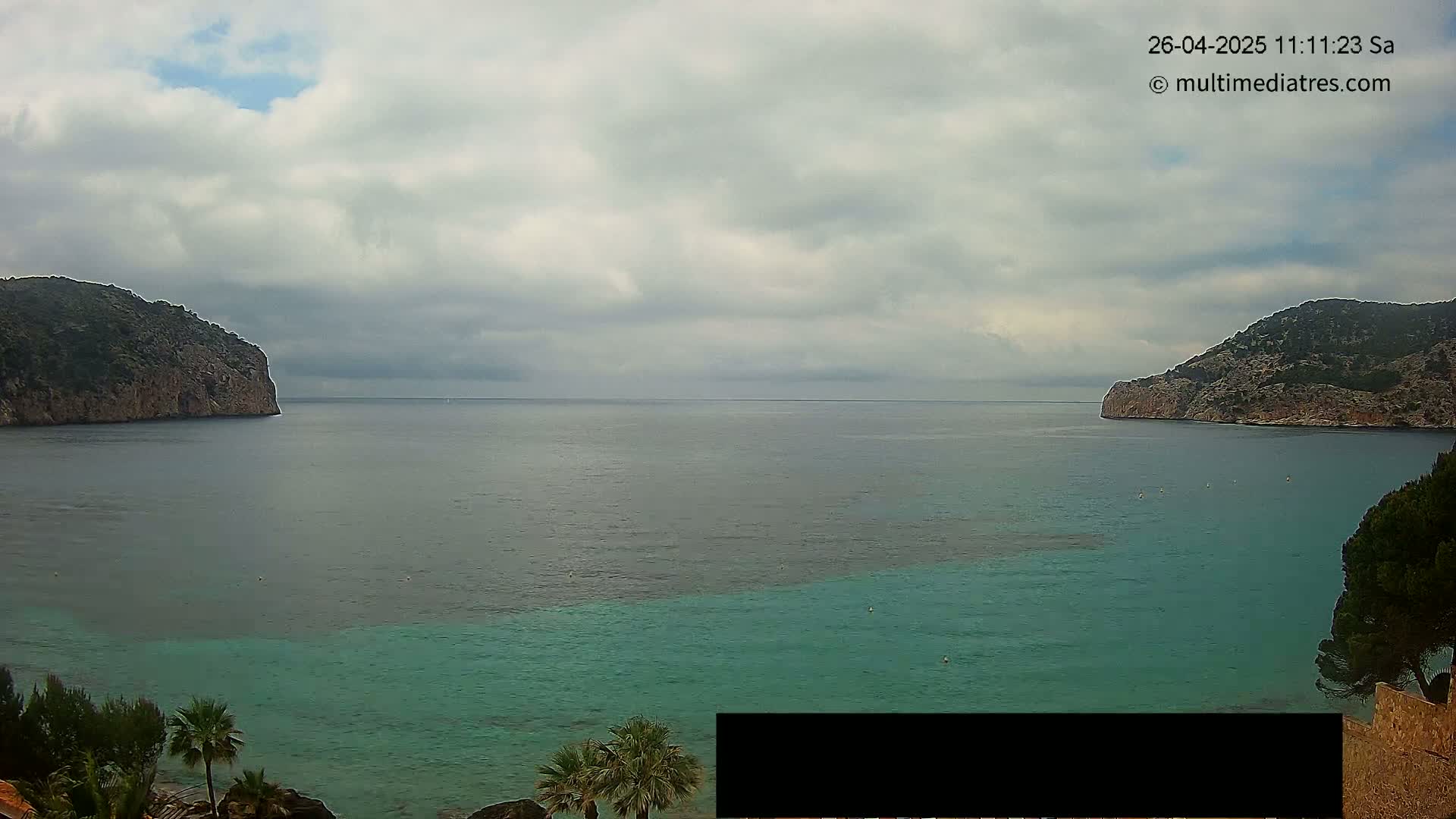 A calm sea with varying shades of teal and blue is nestled between two rocky, green-covered hills under a cloudy sky.