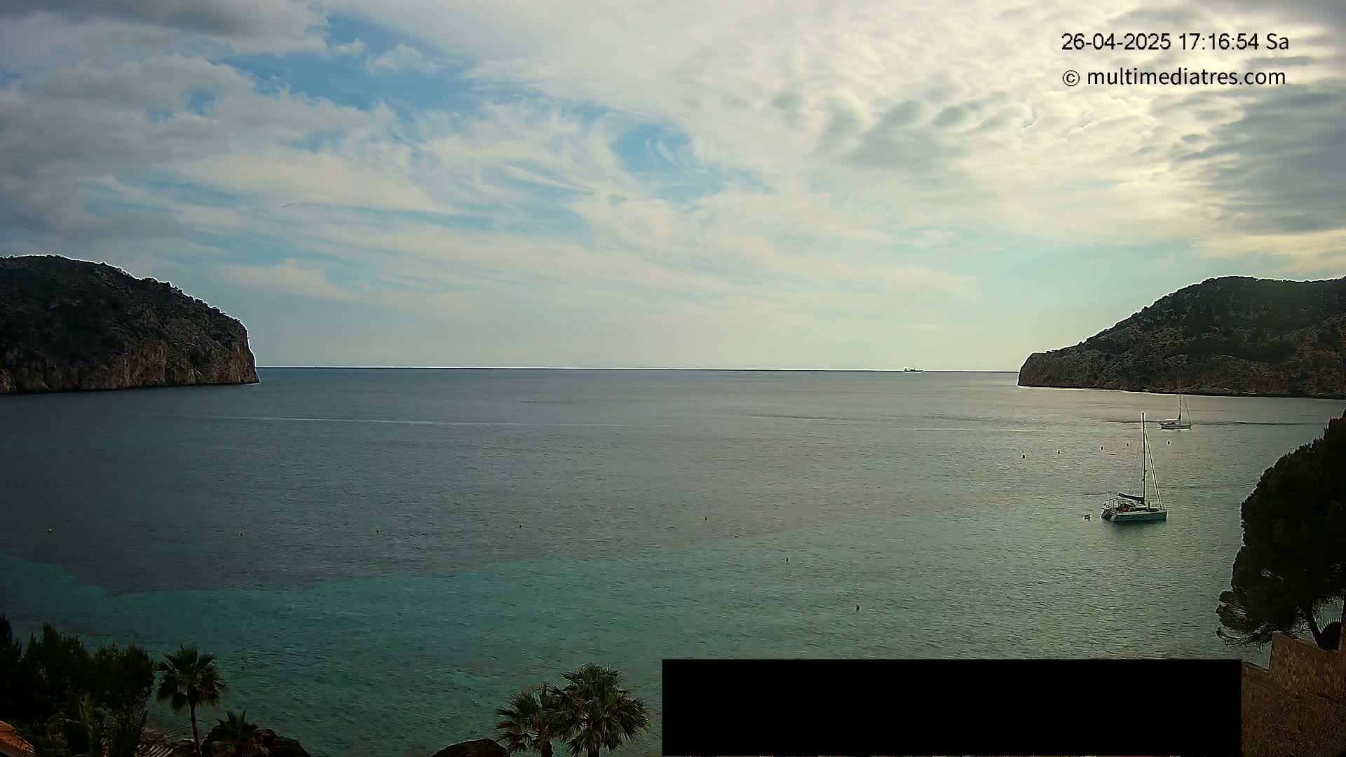 A partly cloudy day reveals a calm bay with two rocky headlands on either side, a sailboat anchored in the water, and vegetation along the shoreline.