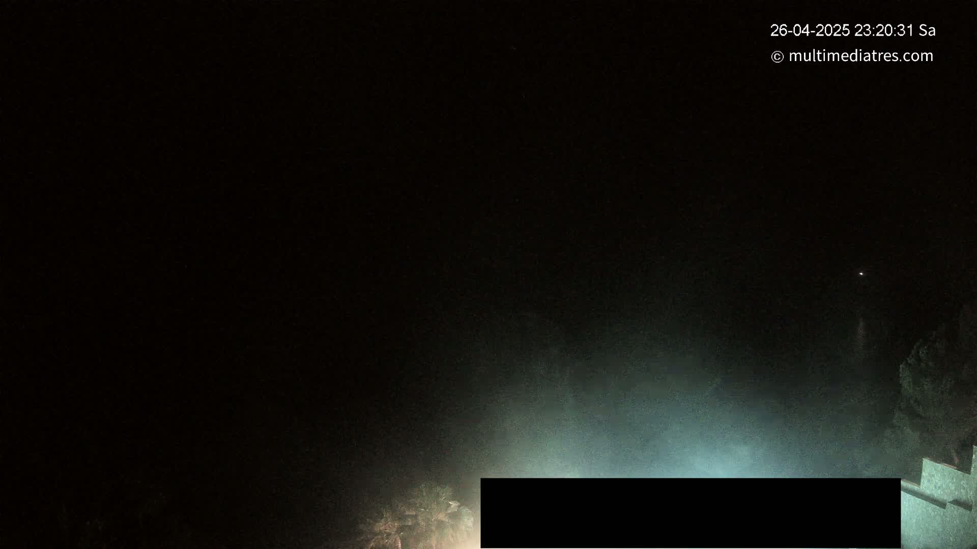 A nighttime outdoor scene shows a hazy, dark sky and a partially visible structure illuminated by a light source.