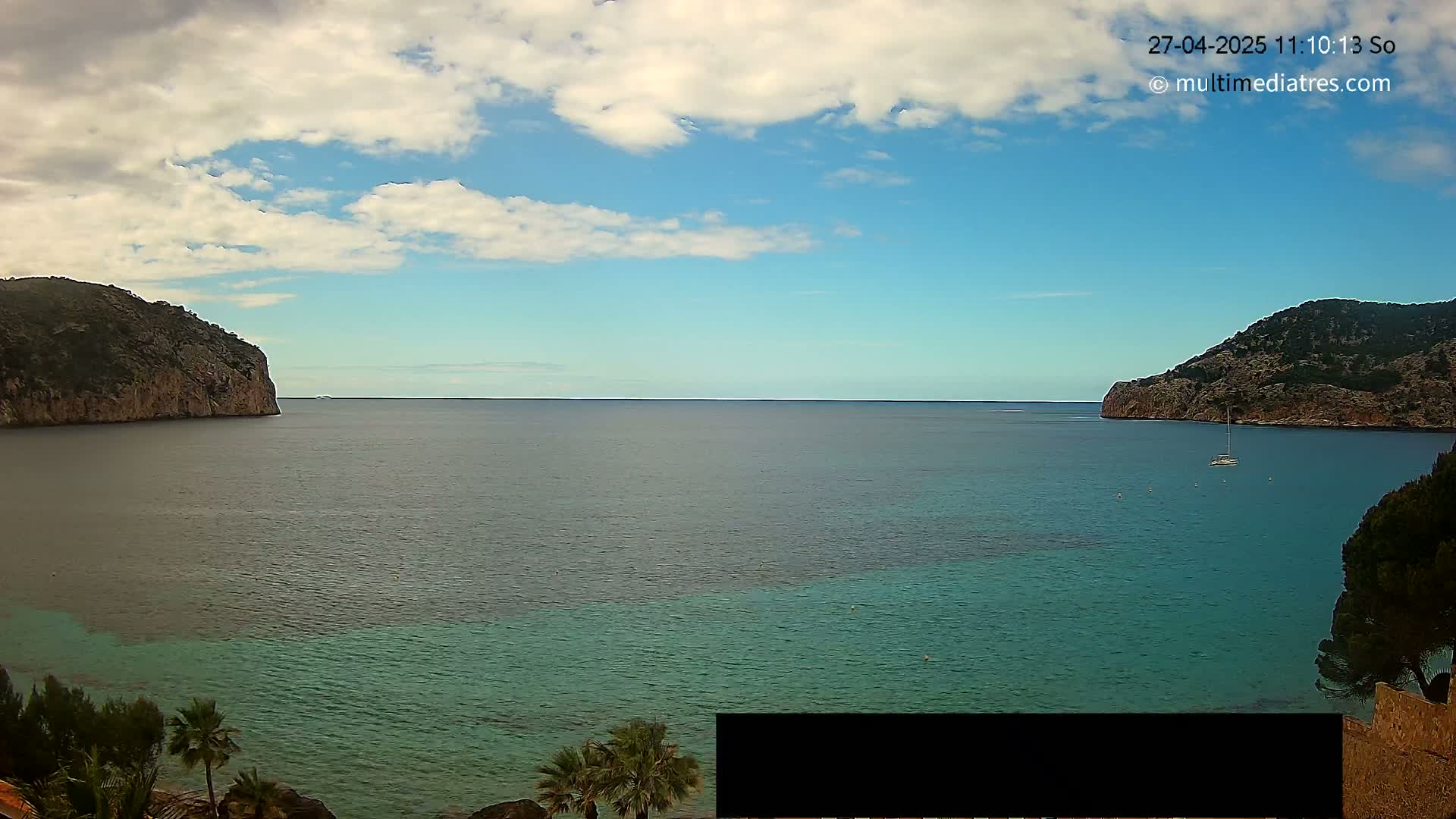 A partly cloudy day shows a calm bay with teal-colored water, nestled between two rocky hillsides, with a sailboat visible in the distance.