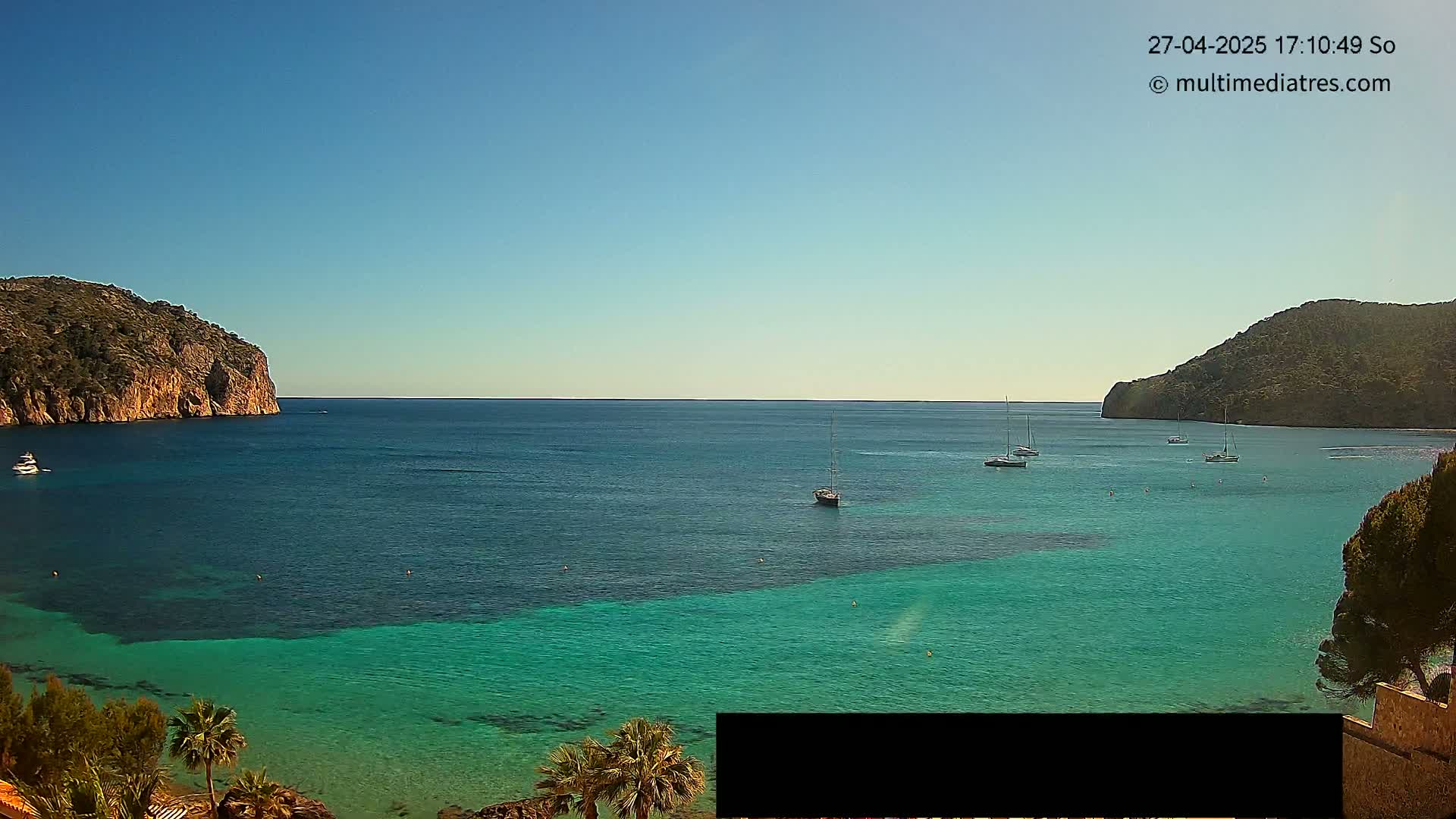 A clear, sunny day shows a calm bay of turquoise water surrounded by rocky hills, with several sailboats at anchor.