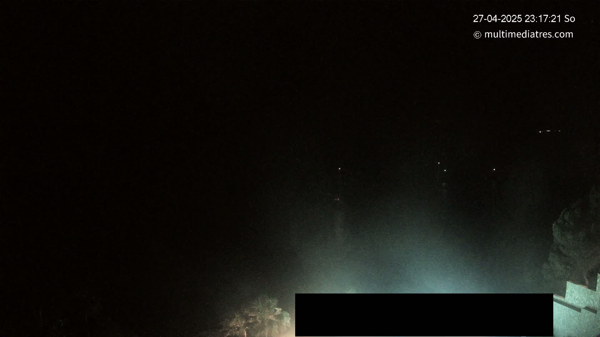 A nighttime view shows a hazy, dark landscape with a few distant lights and some vegetation in the bottom right corner.