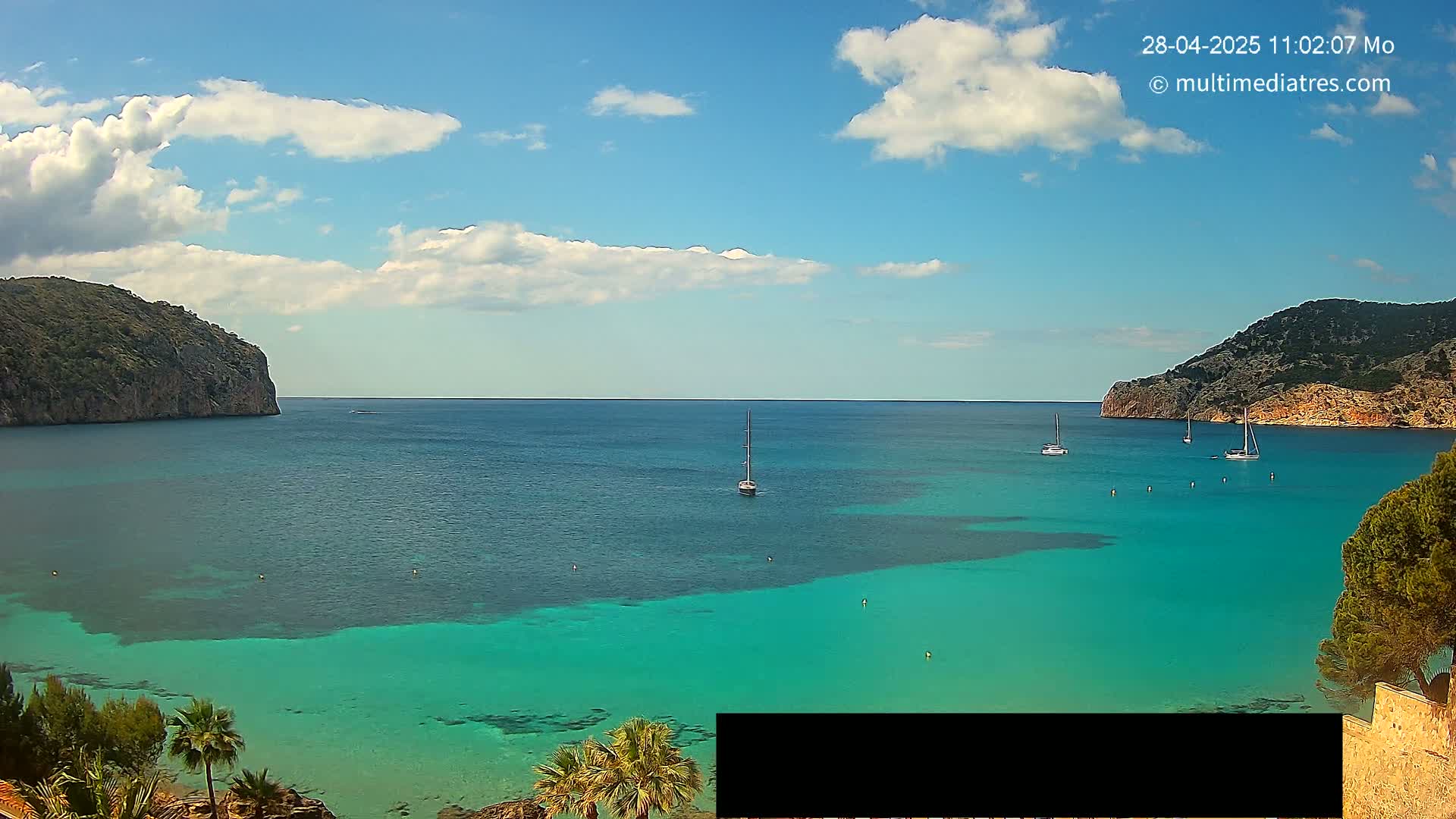 A partly cloudy sky overlooks a calm, turquoise bay surrounded by rocky hills, with several sailboats scattered across the water.