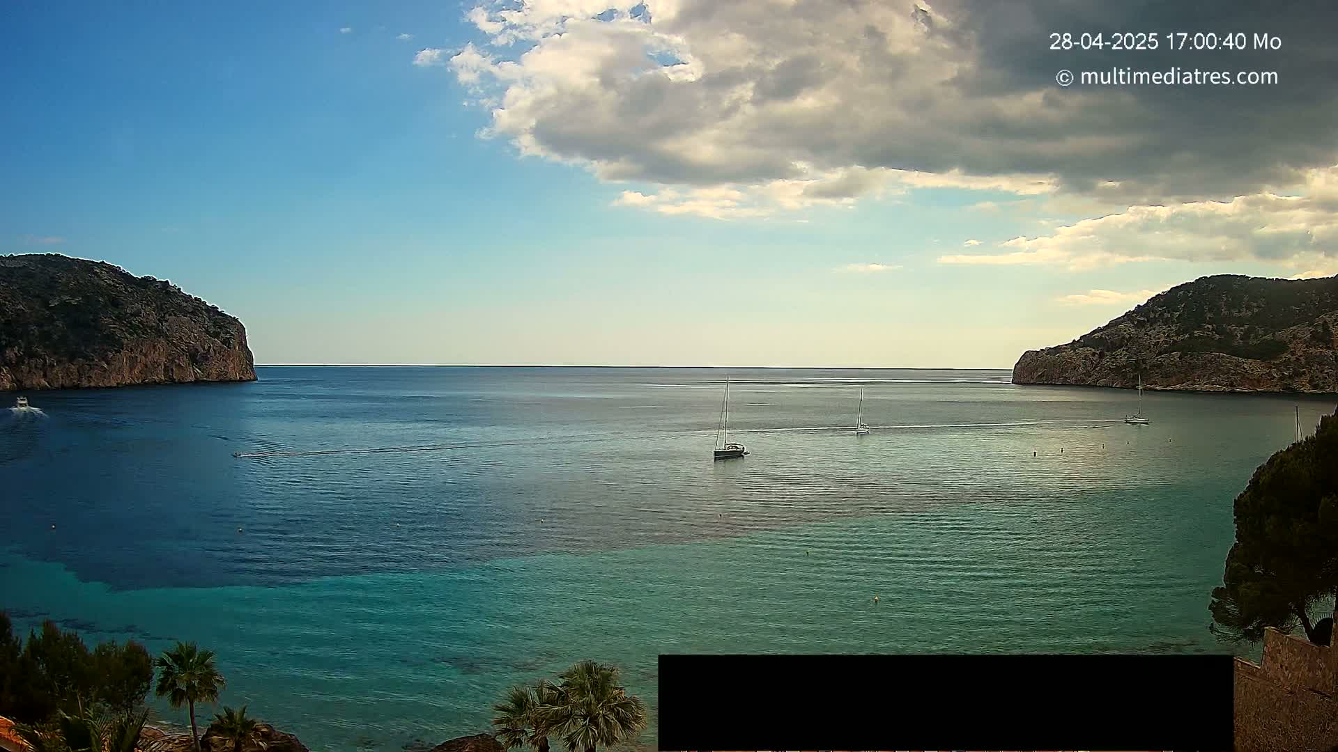A partly cloudy day shows a calm bay of teal and dark blue water nestled between two rocky cliffs, with several sailboats visible on the water.