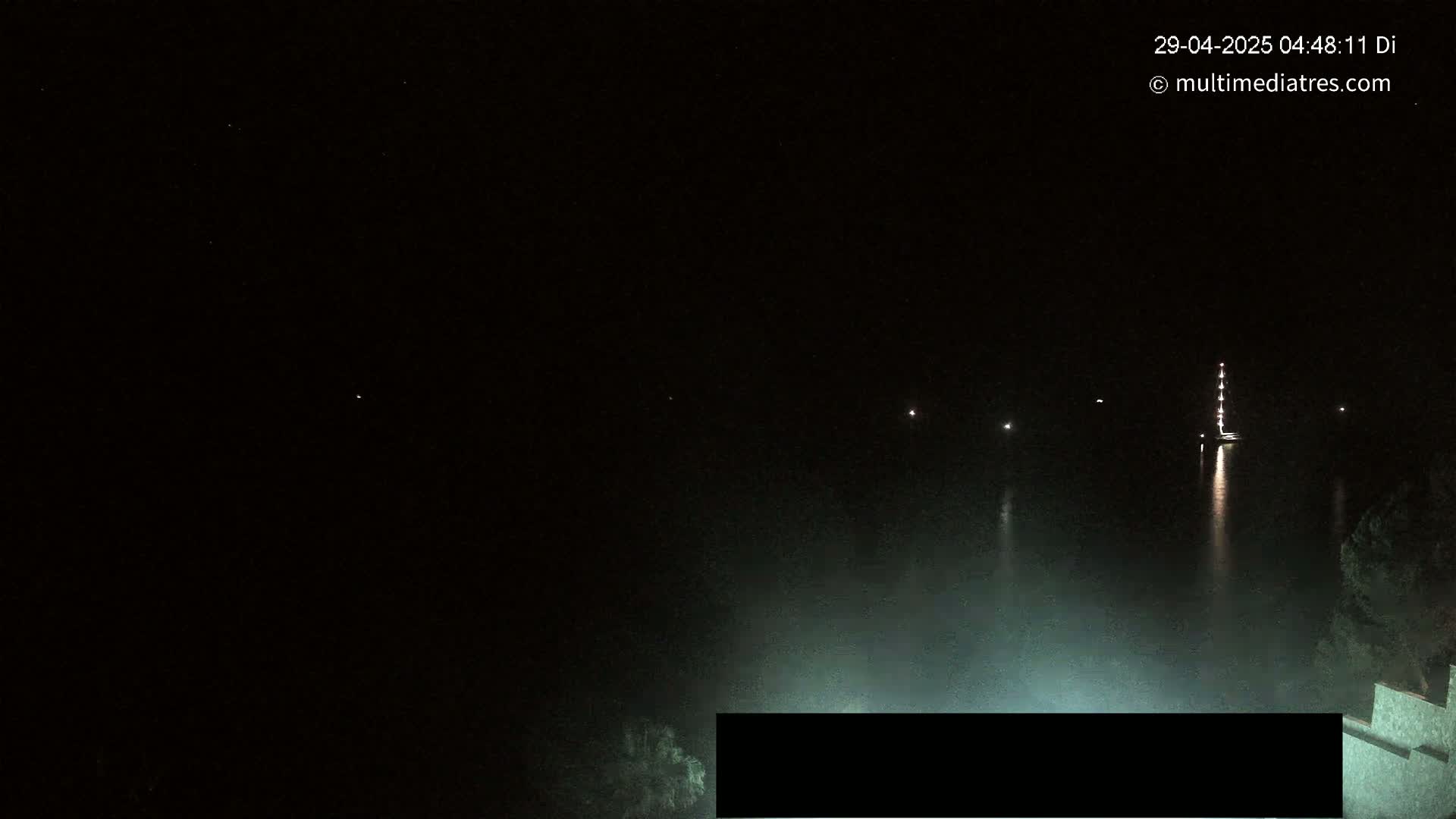 A dark nighttime scene shows a body of water with several small lights and a larger, lit vessel in the distance, partially obscured by a light fog.