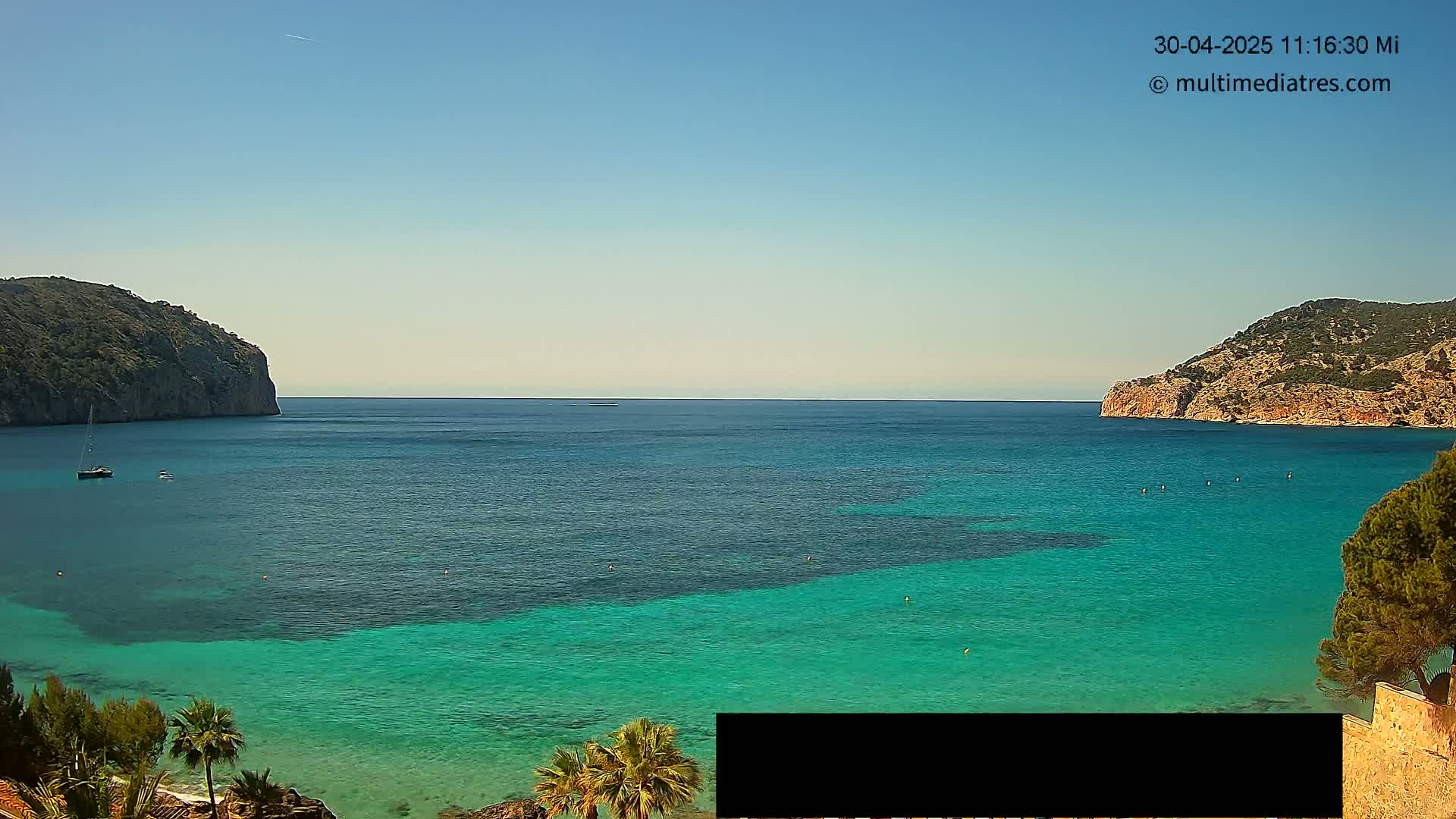 A clear, sunny day reveals a picturesque bay with calm, turquoise water nestled between two rocky hills, a few boats visible on the water's surface.
