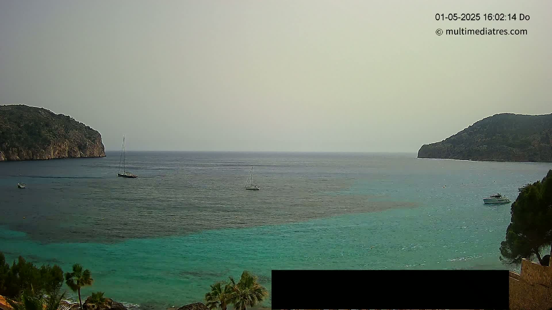 A calm, hazy day reveals a picturesque bay with teal water, several sailboats, and two rocky headlands flanking the tranquil scene.
