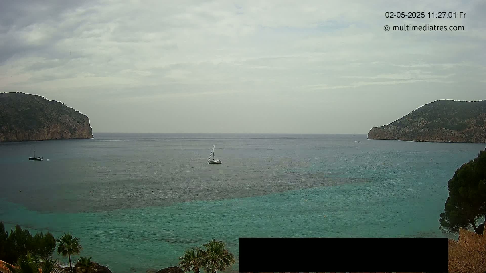 A sailboat floats in a calm, overcast bay between two dark, rocky hillsides.