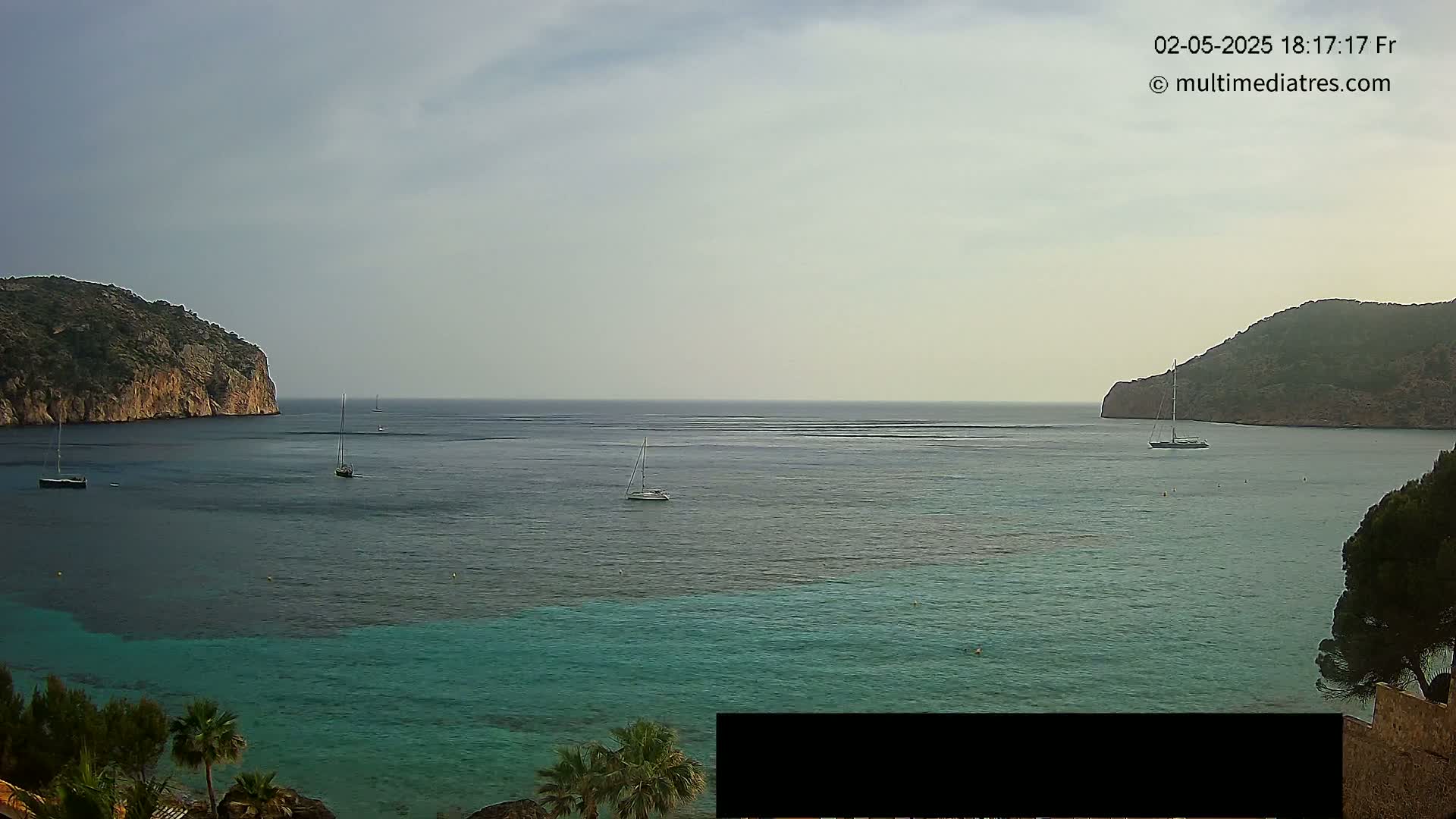 A calm bay with turquoise water is nestled between two rocky hills, with several sailboats scattered across the surface under an overcast sky.