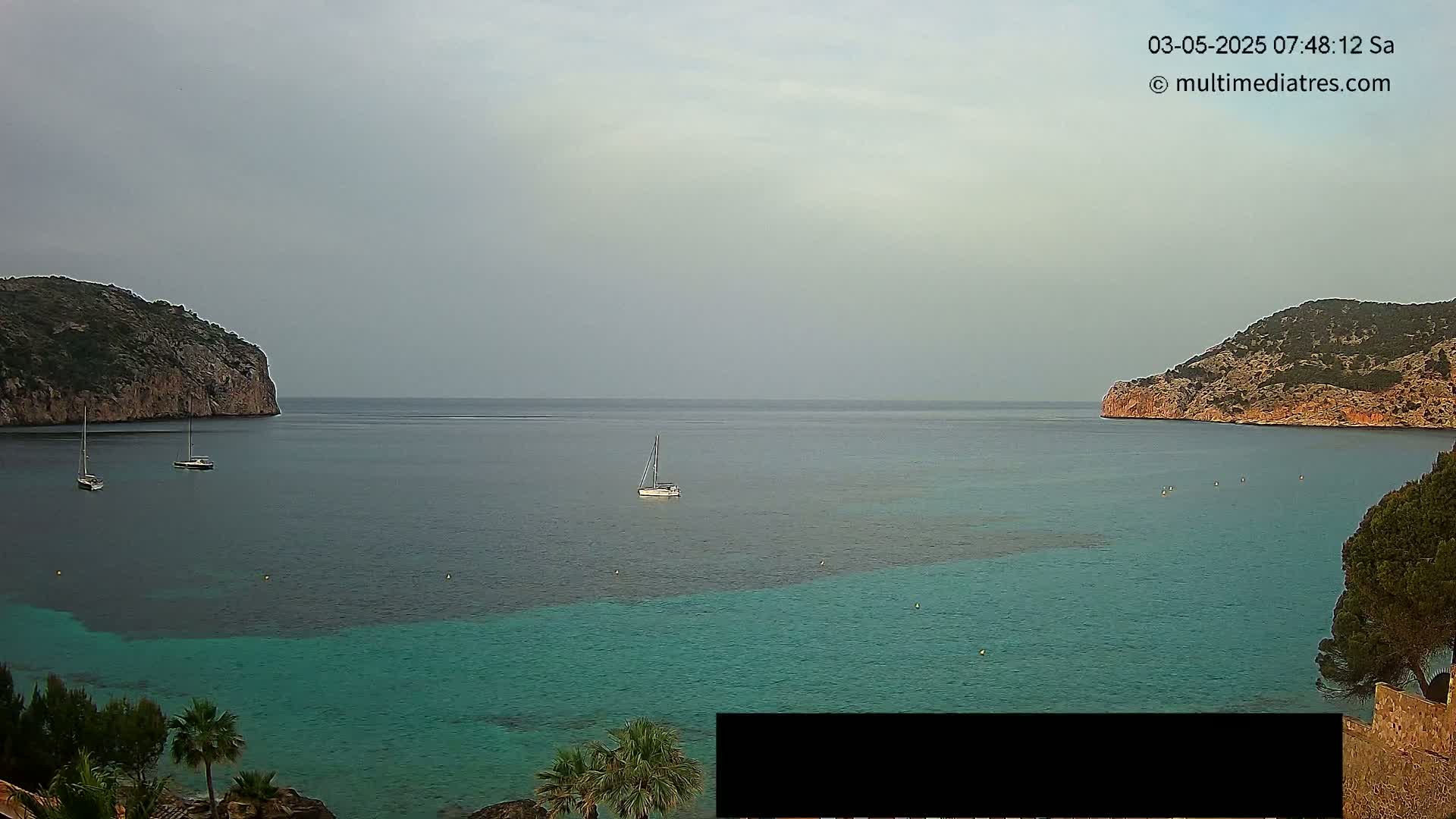 A tranquil cove with calm, teal water is nestled between two rocky hills under an overcast sky; three sailboats are visible in the bay.