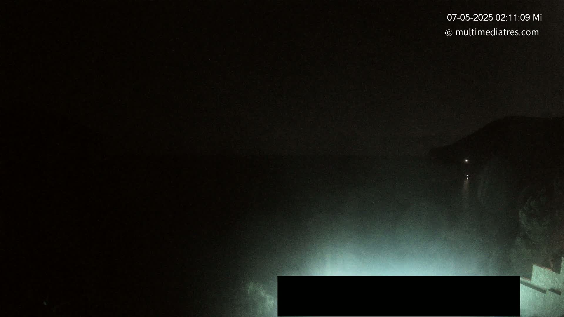 A dark, nighttime scene shows a body of water under a mostly clear sky with a dimly lit shoreline and faint lights in the distance.