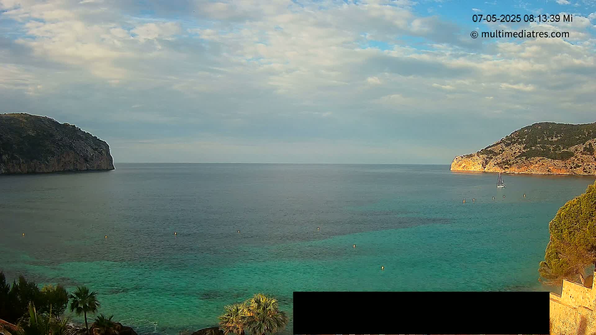 A partly cloudy sky overlooks a calm bay of teal water between two rocky hills, with a sailboat visible in the distance.