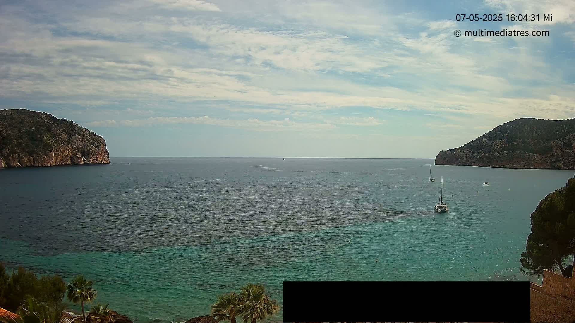 A partly cloudy sky overlooks a calm, teal-colored bay nestled between two rocky, tree-covered hills, with several sailboats visible on the water.