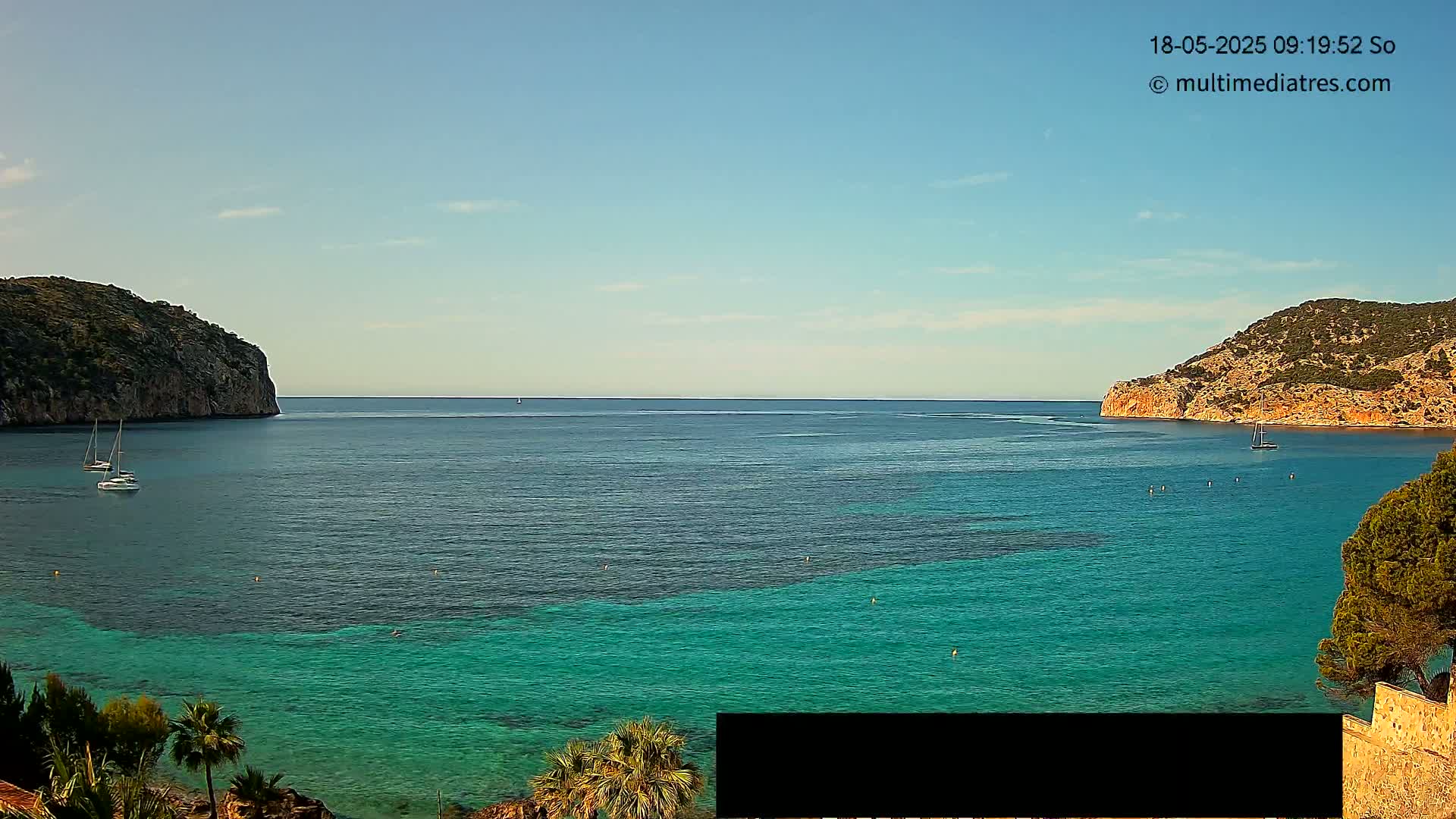 A calm, sunny day shows a turquoise bay nestled between two rocky hills, with a few sailboats visible on the water.