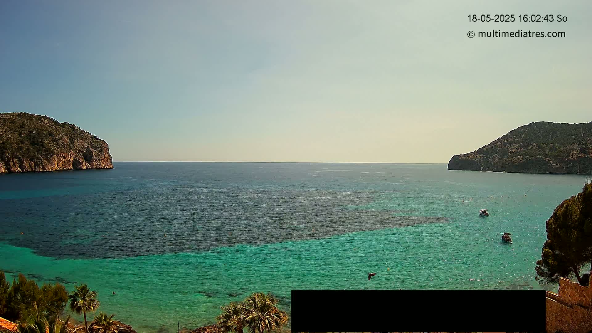 A calm, sunny day reveals a cove of clear, turquoise water nestled between two rocky hillsides, with a few small boats visible on the water.