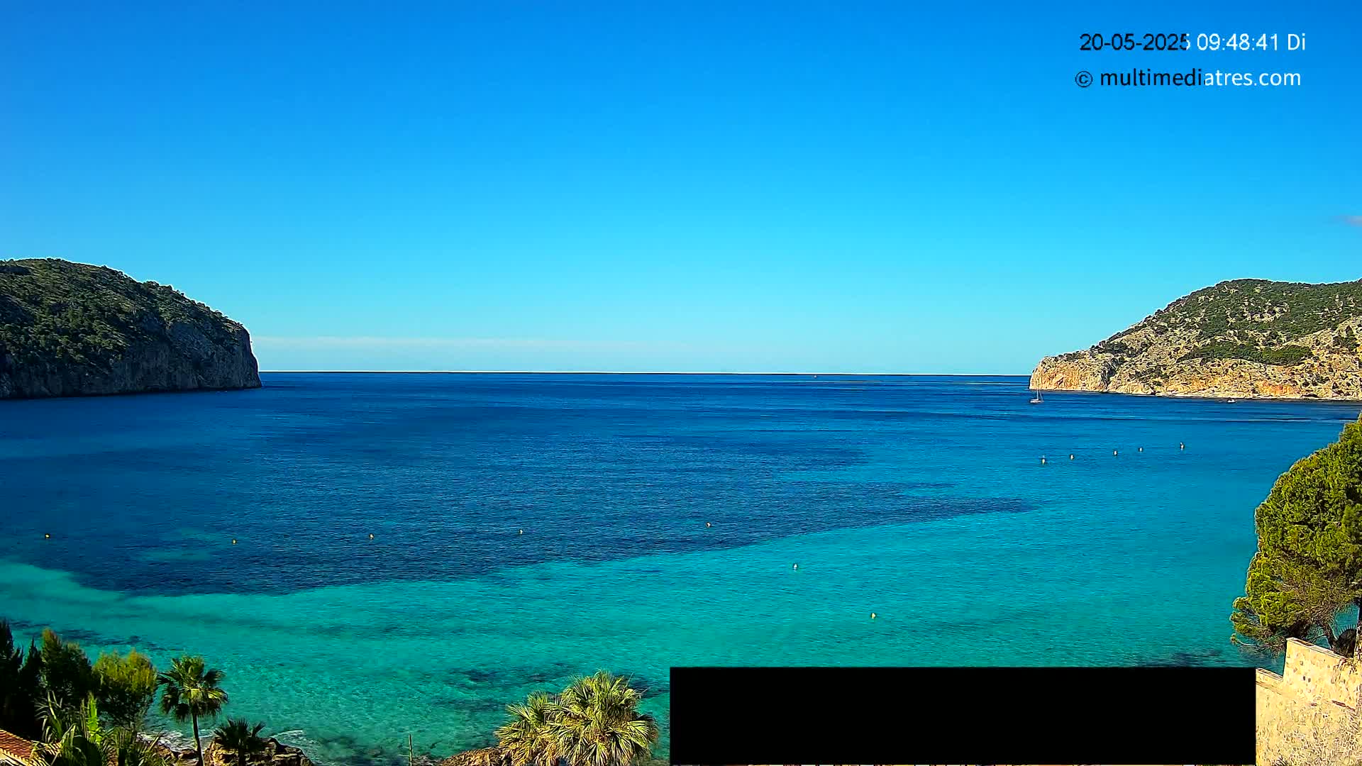 A clear blue sky overlooks a calm, bright turquoise sea nestled between two verdant hills.