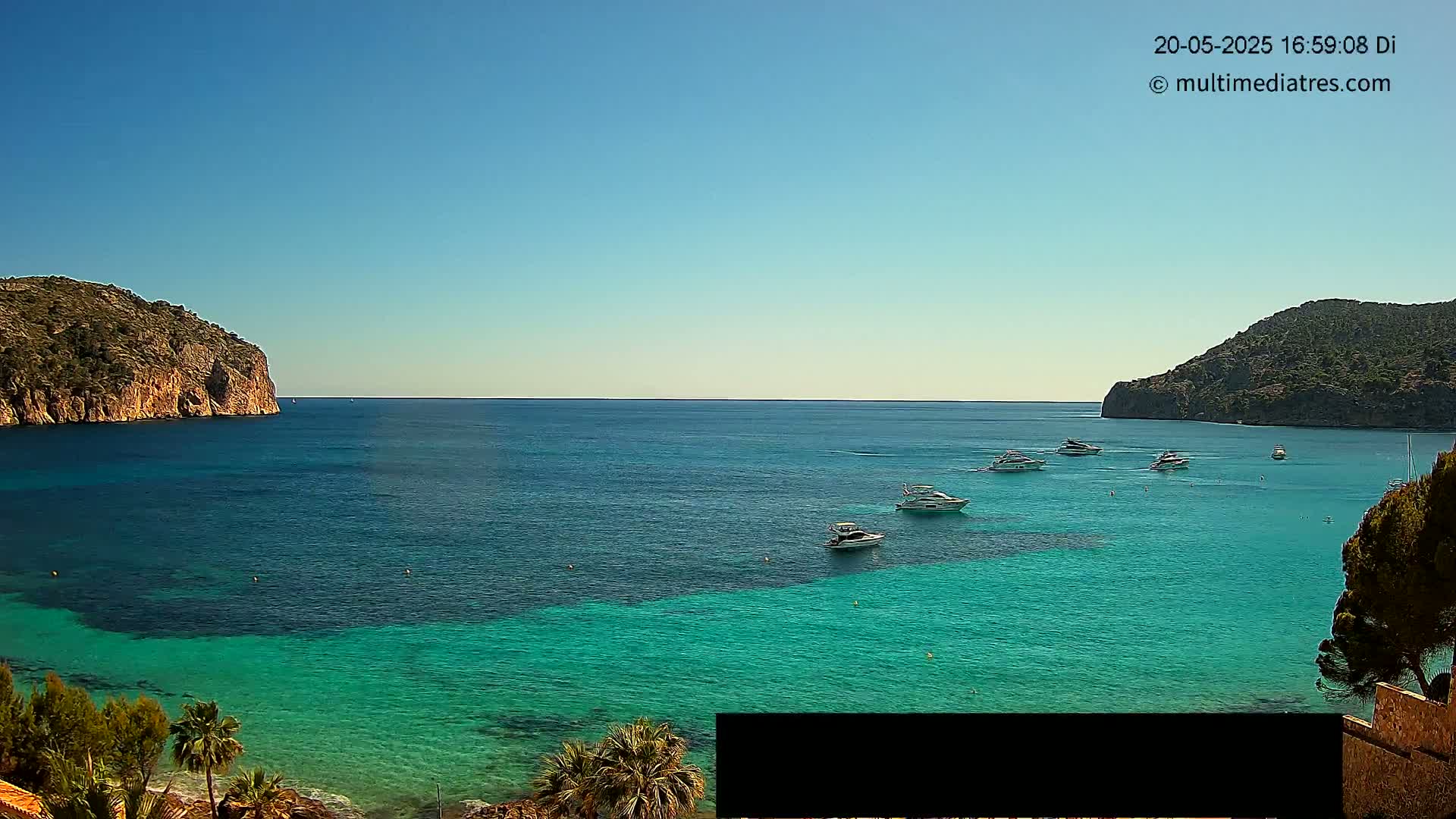 A clear, sunny day reveals a calm bay of vibrant turquoise water, with several boats anchored near rocky headlands.