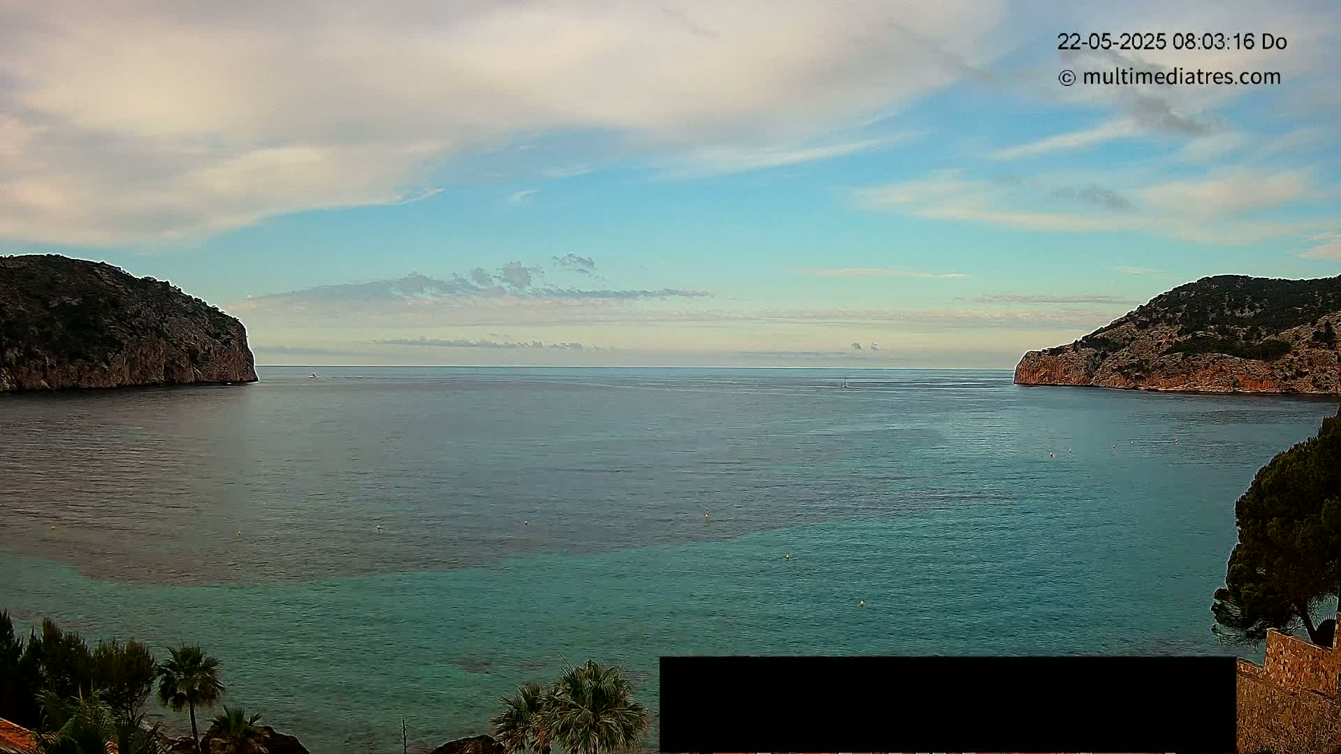 A calm sea with partly cloudy skies is nestled between two rocky hillsides, with some vegetation visible at the bottom.