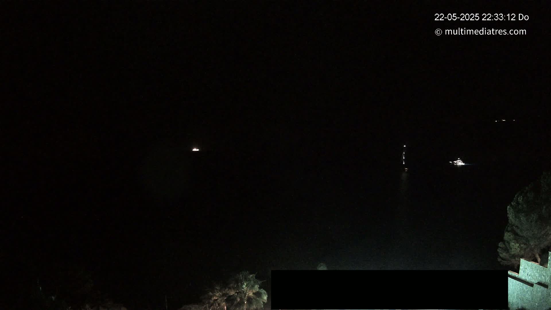At night, a dark body of water is visible with several small lights from boats or ships in the distance, and some vegetation is seen on the lower right corner of the image.