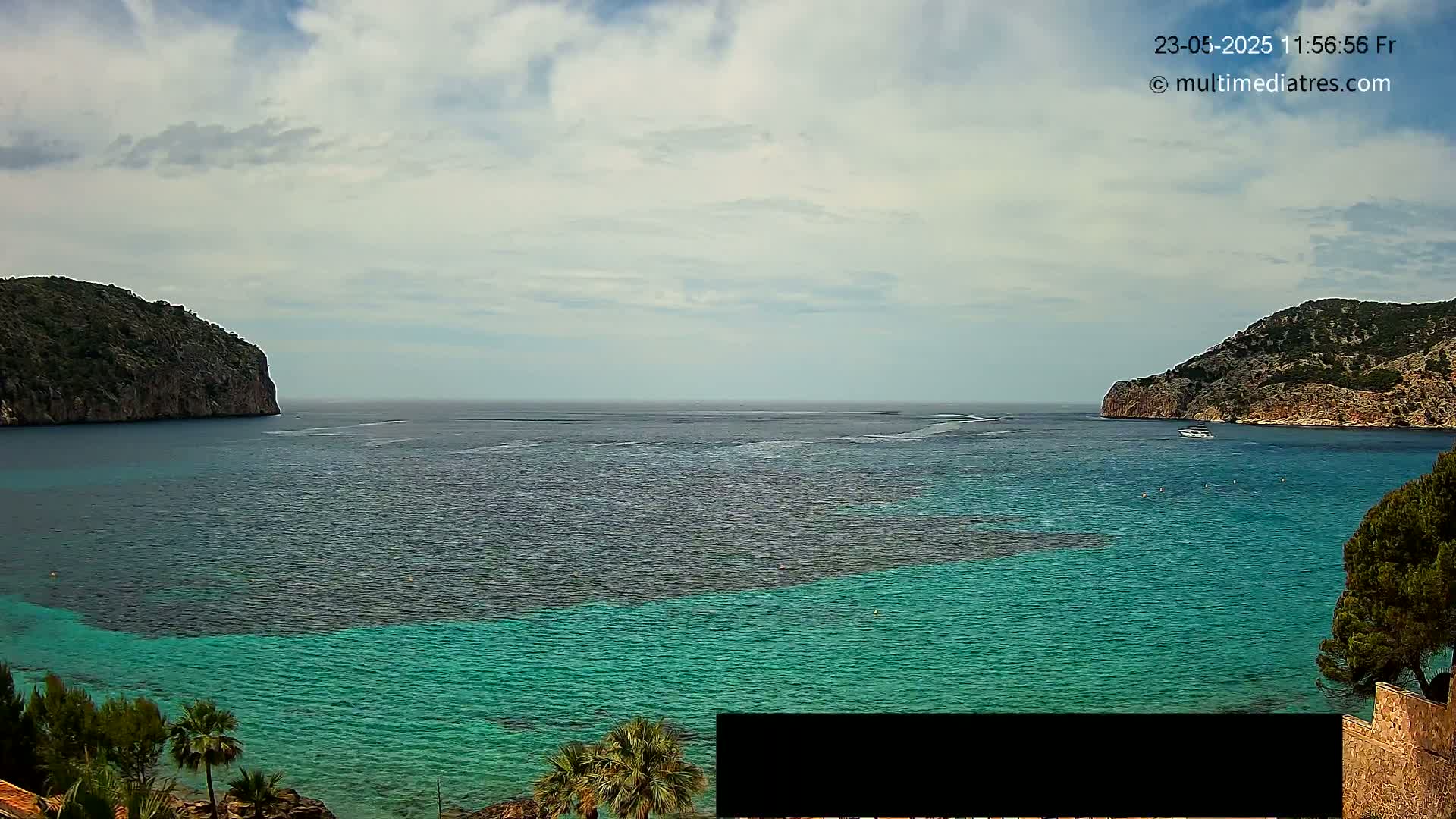 A partly cloudy day reveals a tranquil turquoise bay nestled between two rocky headlands, with a boat visible on the calm water.